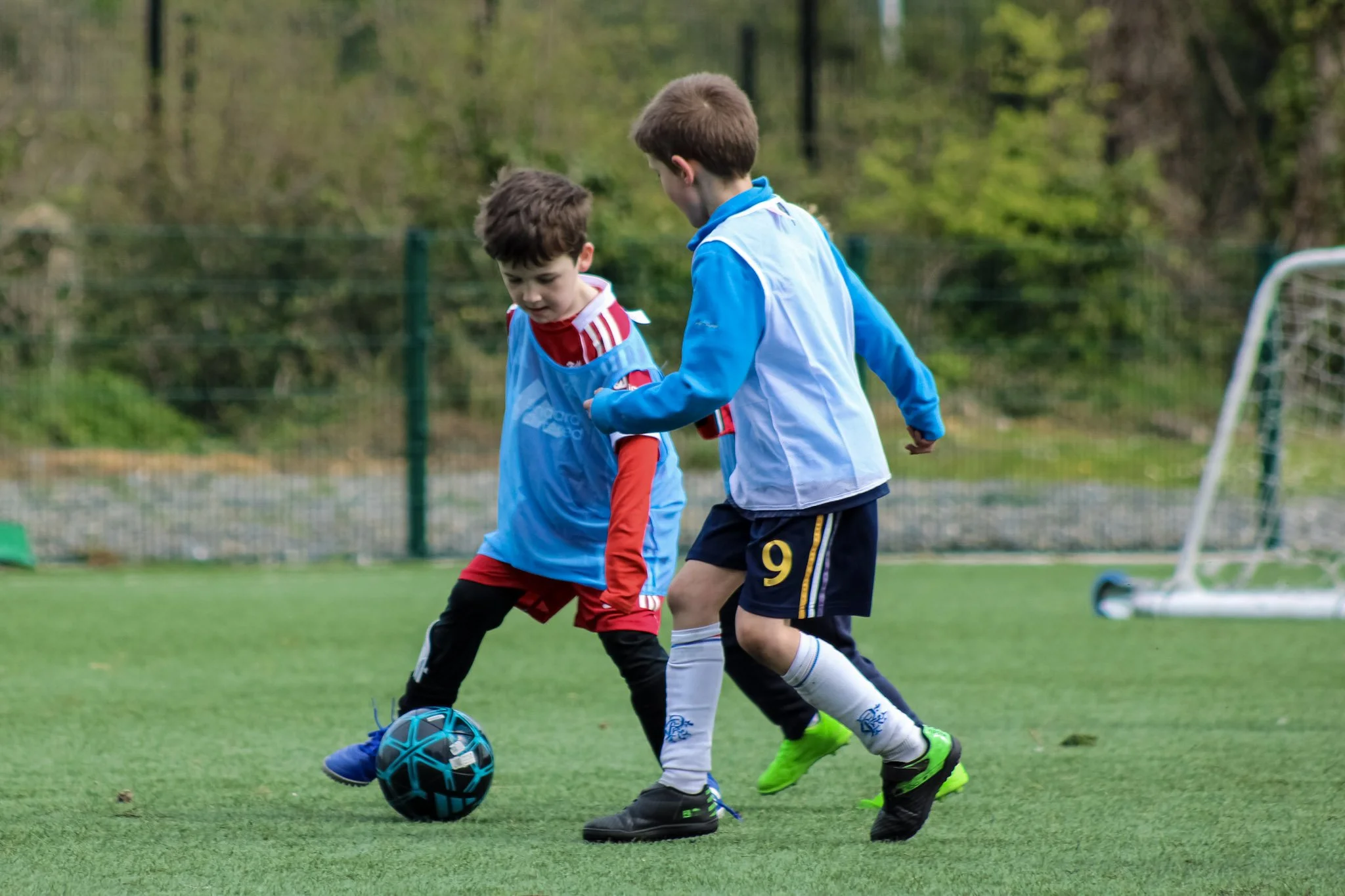 Two boys playing soccer on a green field, with trees and a fence in the background. One boy in a blue and white jersey is dribbling the ball while the other boy in a red and blue jersey is trying to take it.