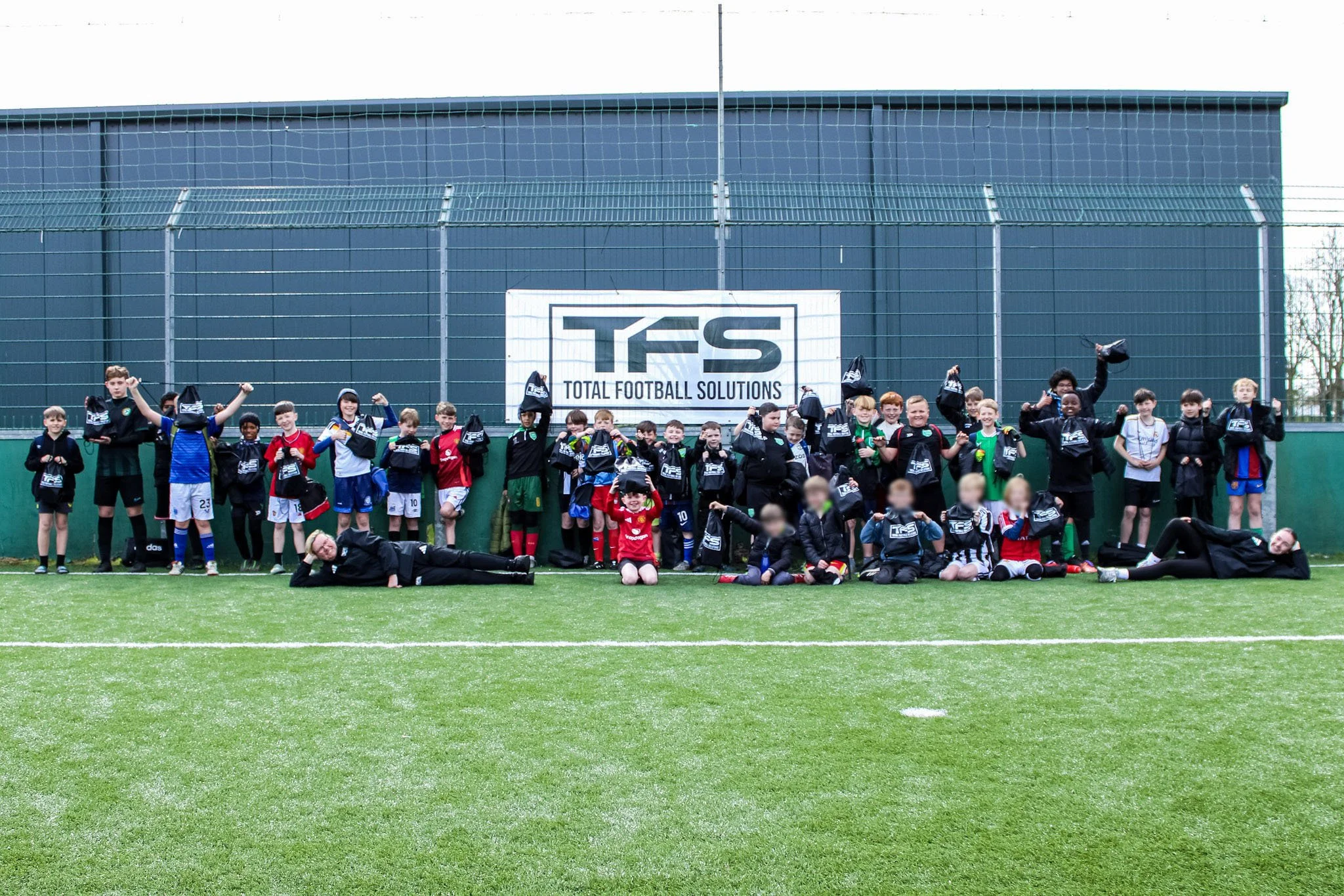 A group of children and teenagers posing for a photo on a football field, with a blue building and a banner reading 'TFS Total Football Solutions' in the background. Some children are holding black drawstring bags.