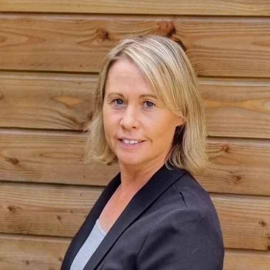 A woman with blonde hair wearing a black blazer and gray top standing in front of a wooden fence.