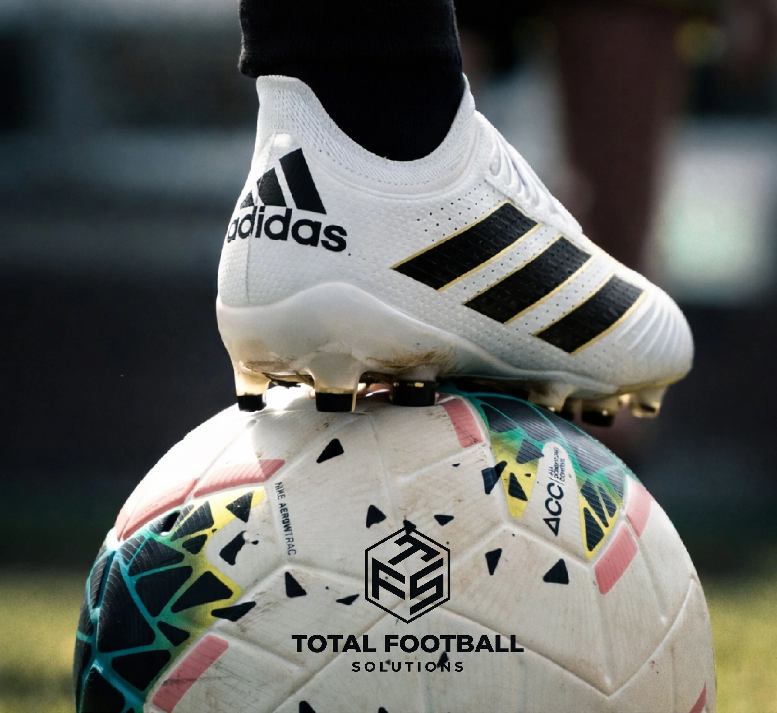 Close-up of a white Adidas football cleat stepping on a colorful soccer ball with the logo 'TOTAL FOOTBALL SOLUTIONS' and other branding on the ball.