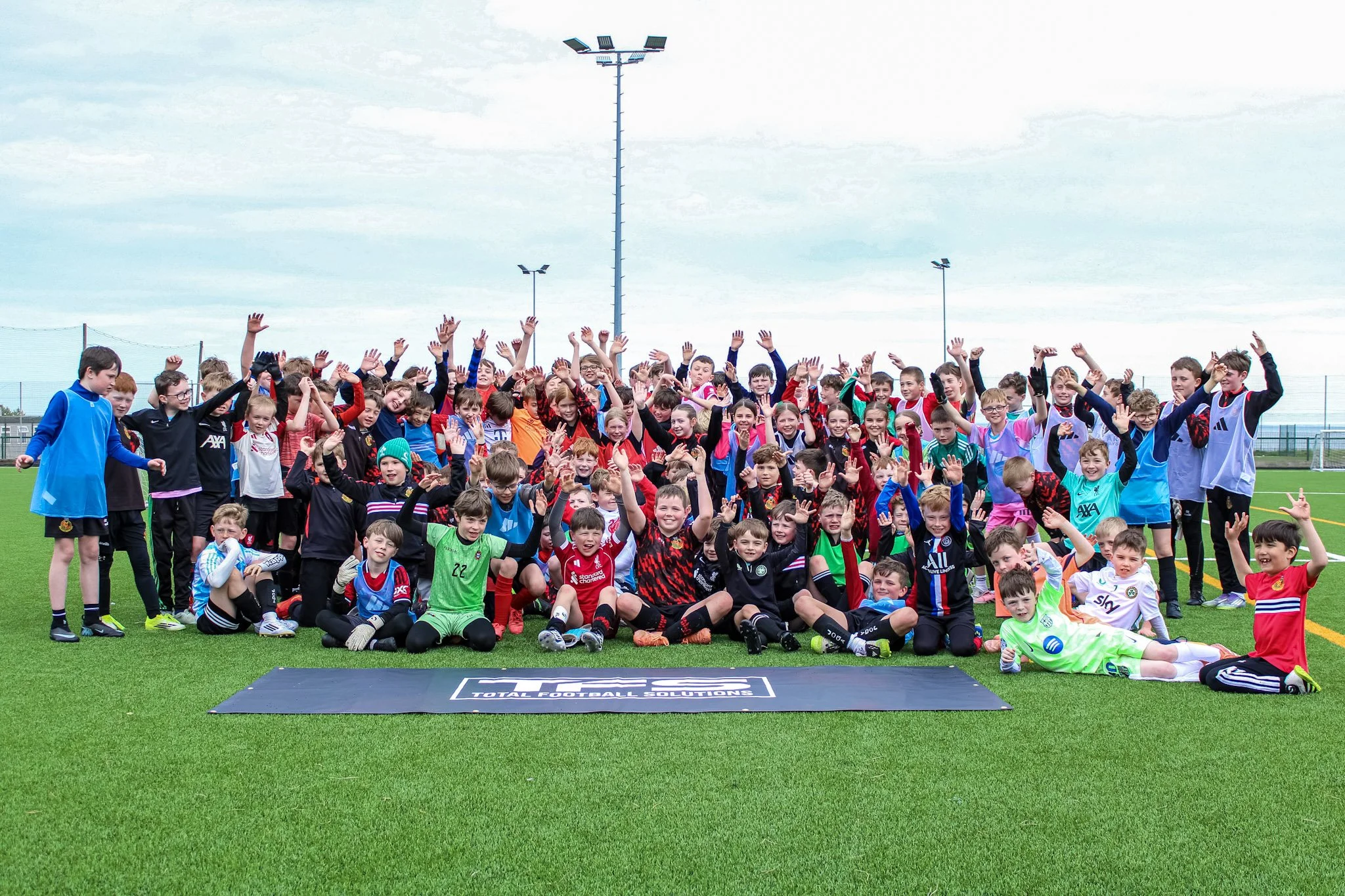 A large group of children and teenagers in sports uniforms, gathered on a sports field for a group photo, with a cloudy sky and tall floodlights in the background.
