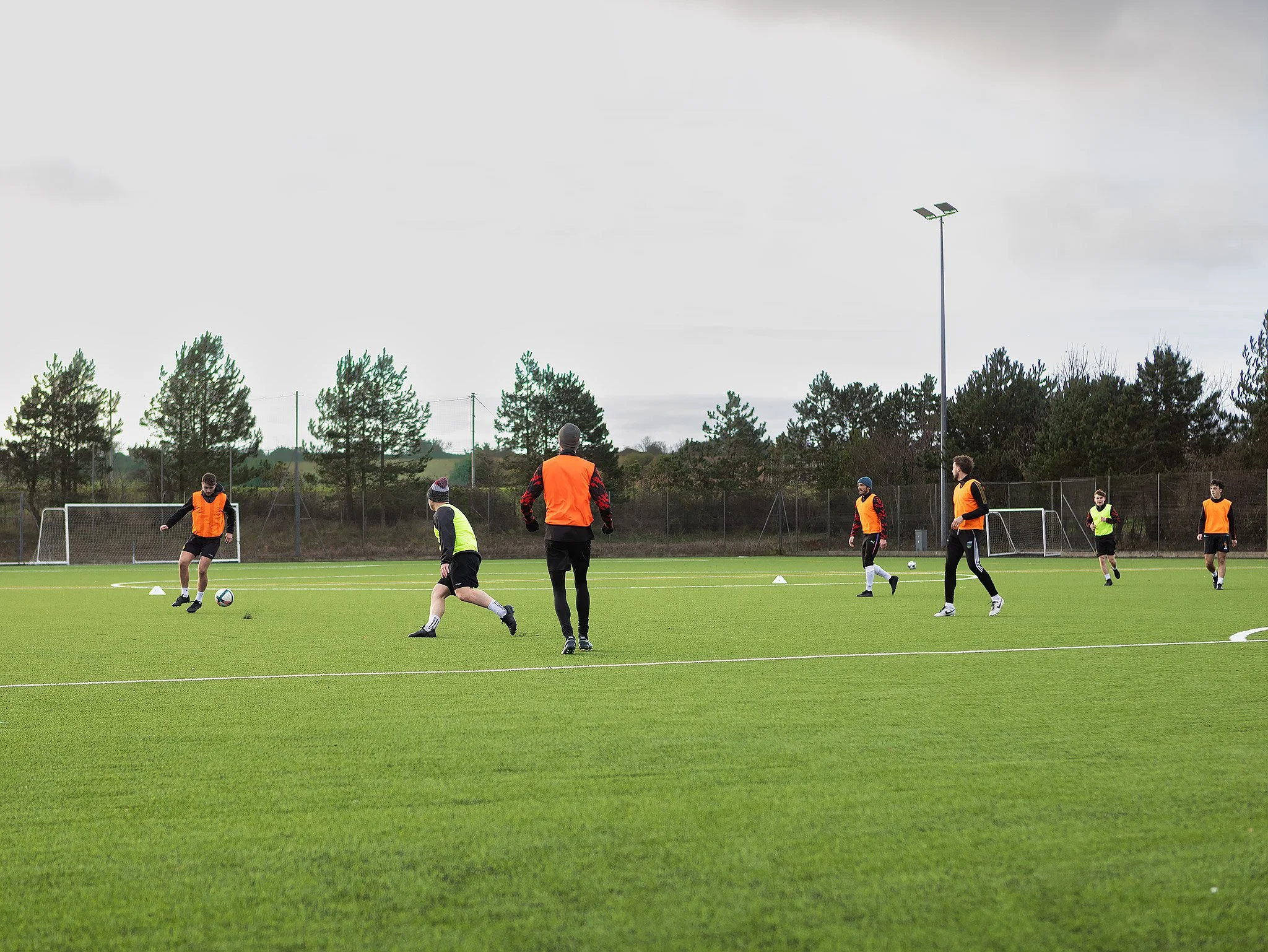 Soccer players practicing on a green field during daytime, some wearing orange pinnies, with goalposts and trees in the background.