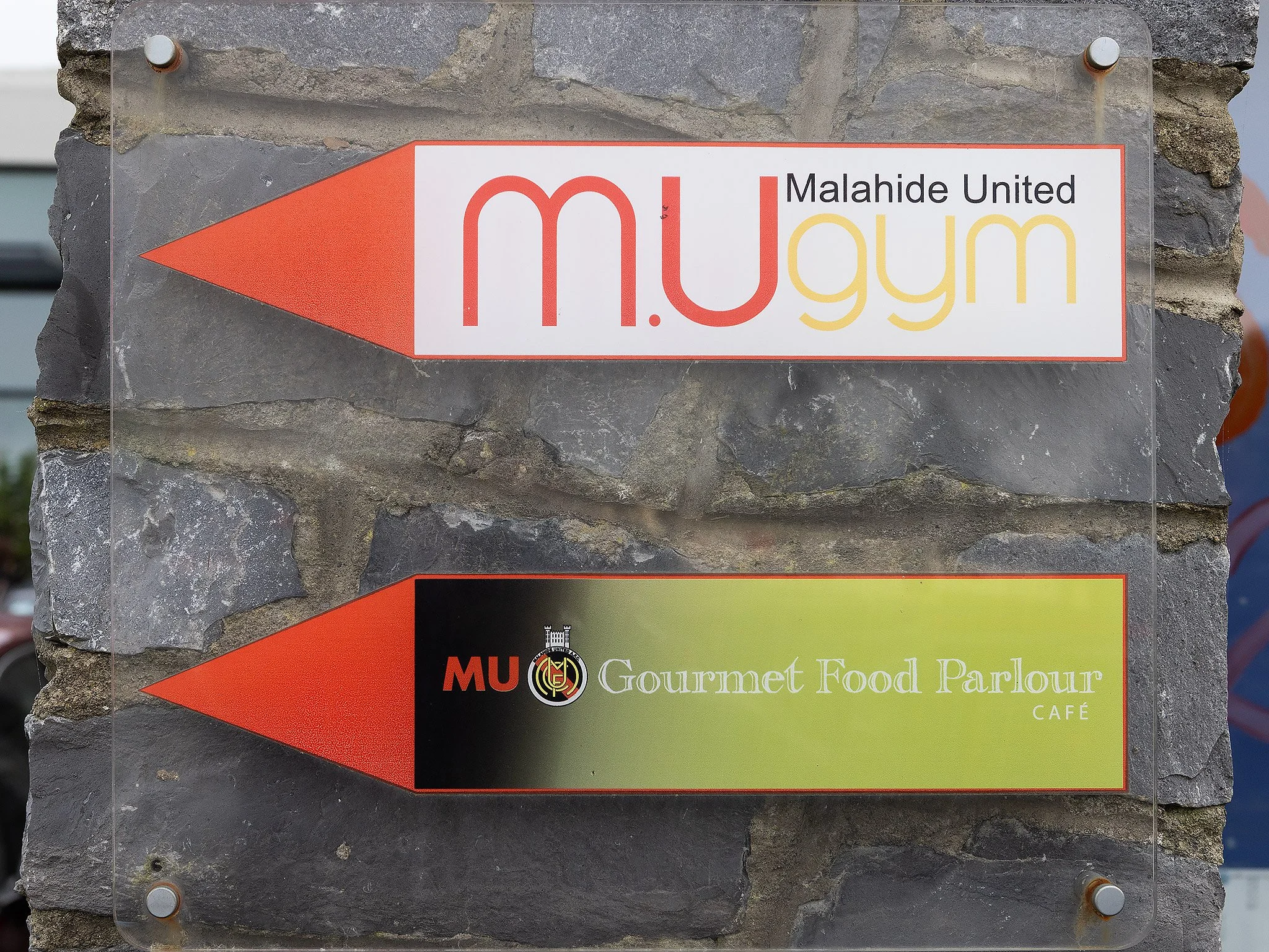 Directional signs pointing to M. U gym and gourmet food parlour on a stone wall.