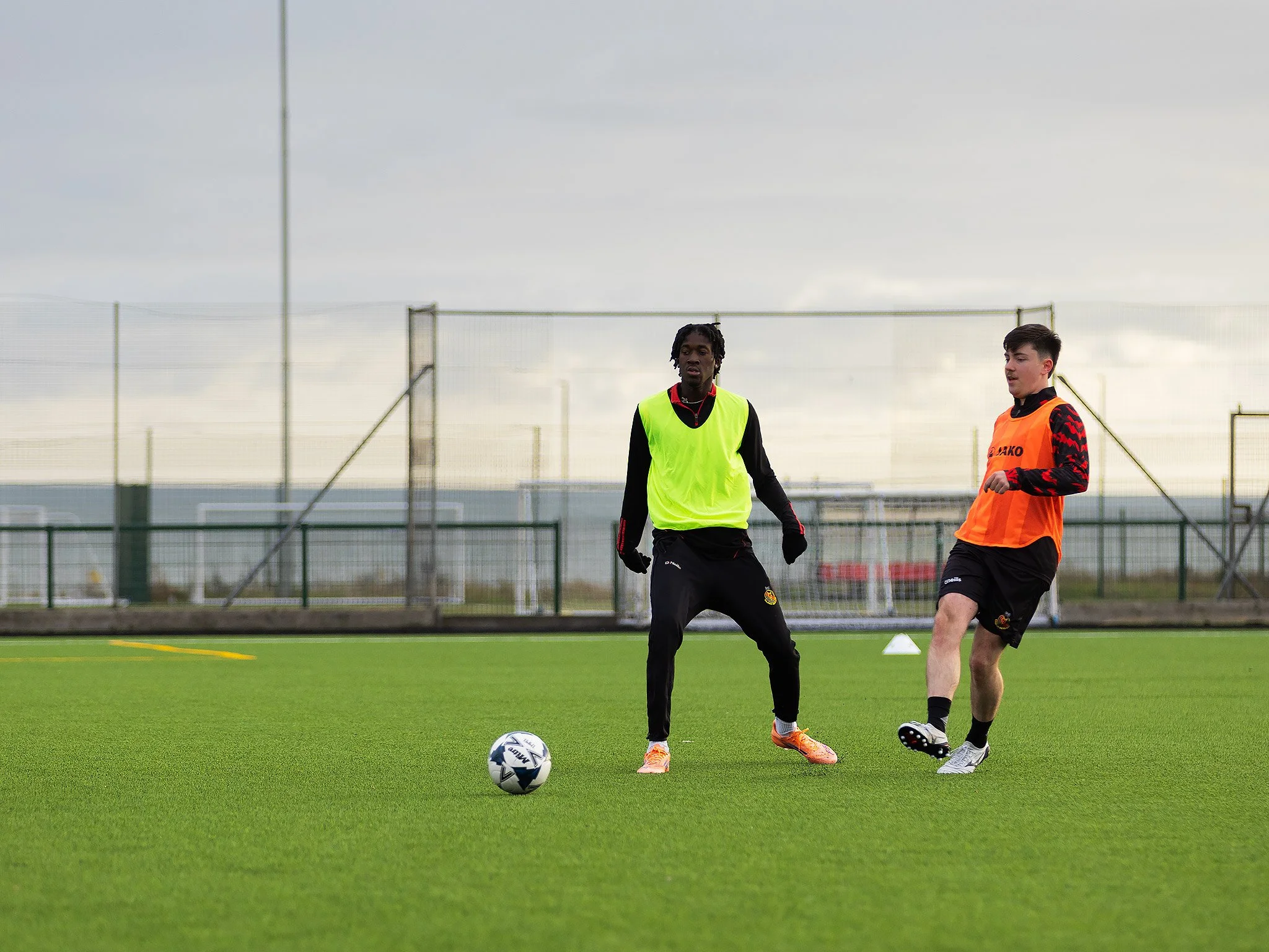 Two young male soccer players practicing on a field with a soccer ball, one wearing a neon green training vest and the other an orange vest, with a fence and goal in the background under a cloudy sky.