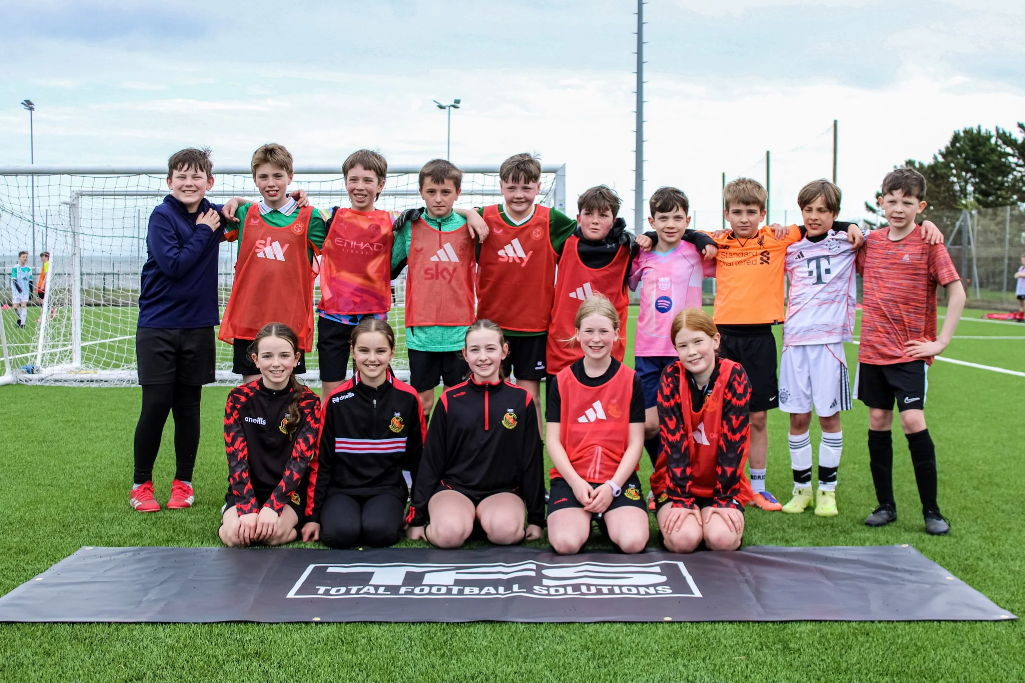 A group of young children and teenagers, some wearing soccer uniforms and others in casual athletic wear, standing together on a soccer field in front of a goalpost, smiling for a photo
