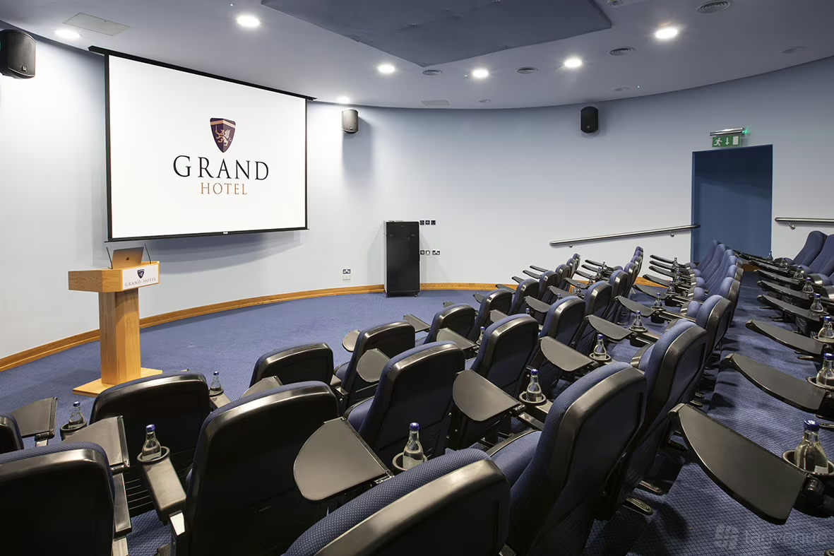 Conference room at Grand Hotel with rows of black chairs, a large screen displaying the hotel logo, and a wooden podium.