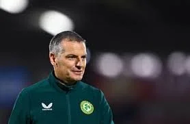 A man with short gray hair and a dark green sports jacket standing outdoors at night with blurred lights in the background.