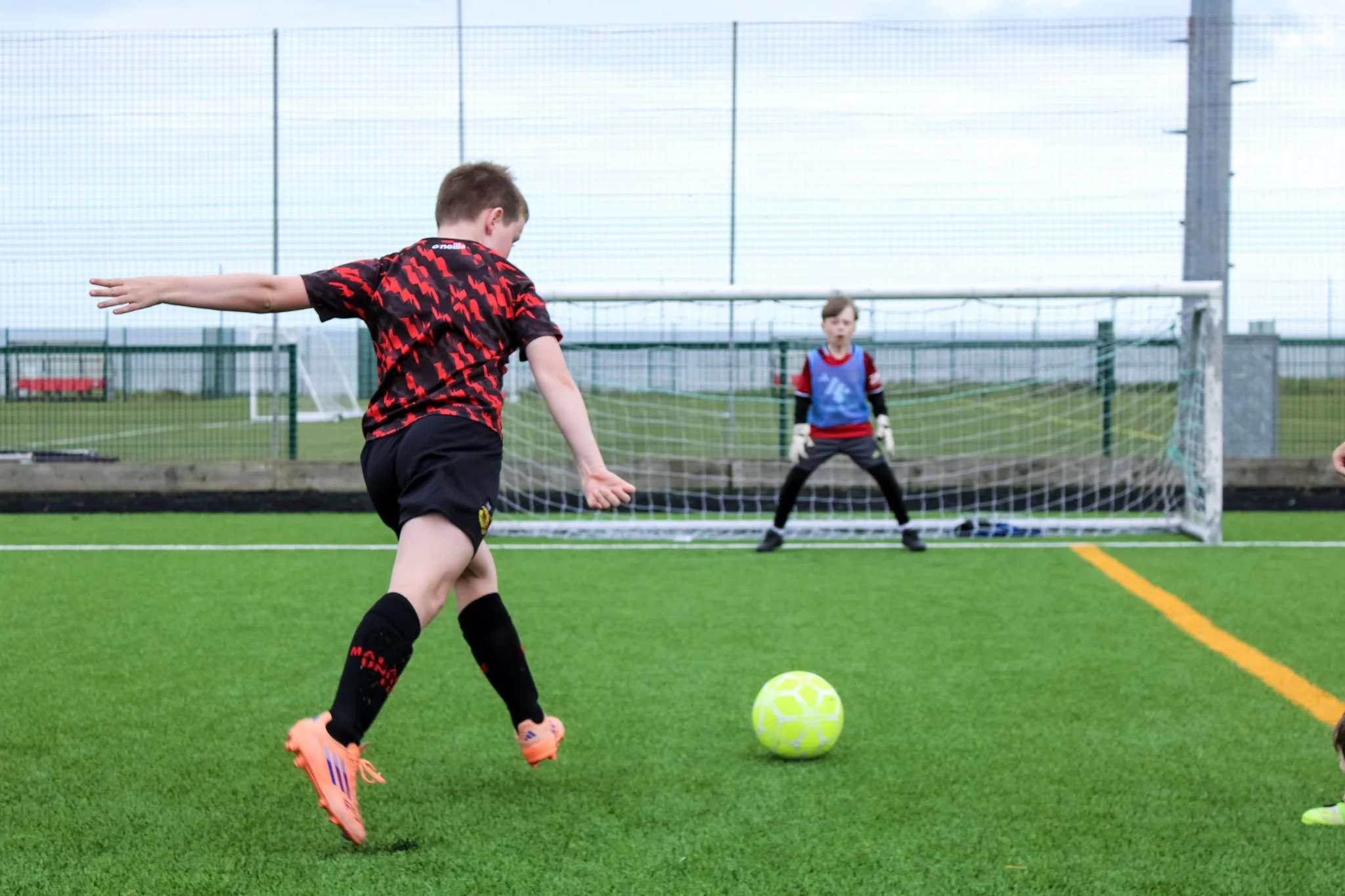 A young boy wearing a black and red soccer jersey, black shorts, and orange soccer cleats kicking a yellow soccer ball on an outdoor field. In the background, another boy in a goalkeeper uniform stands in front of a goal with a net, on a grassy sports field surrounded by a fence.