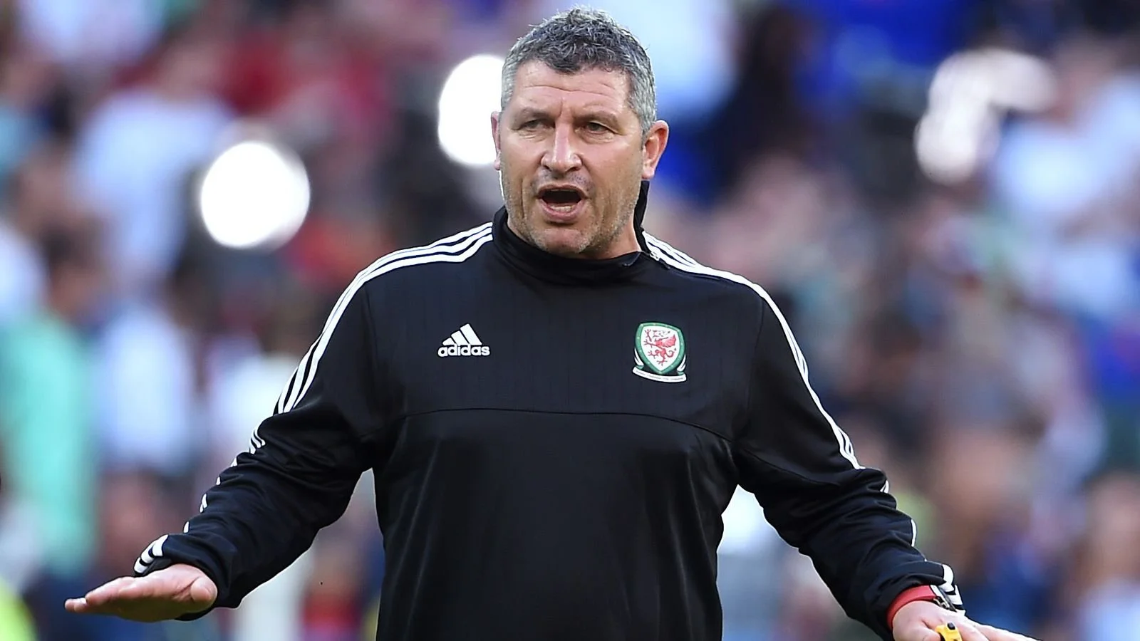 A man in a black sports jacket with white stripes and a badge, speaking or gesturing on a sports field with a crowd in the background.