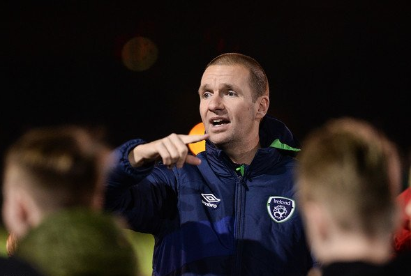 A man in a blue sports jacket talking and gesturing during a sports event, surrounded by children.