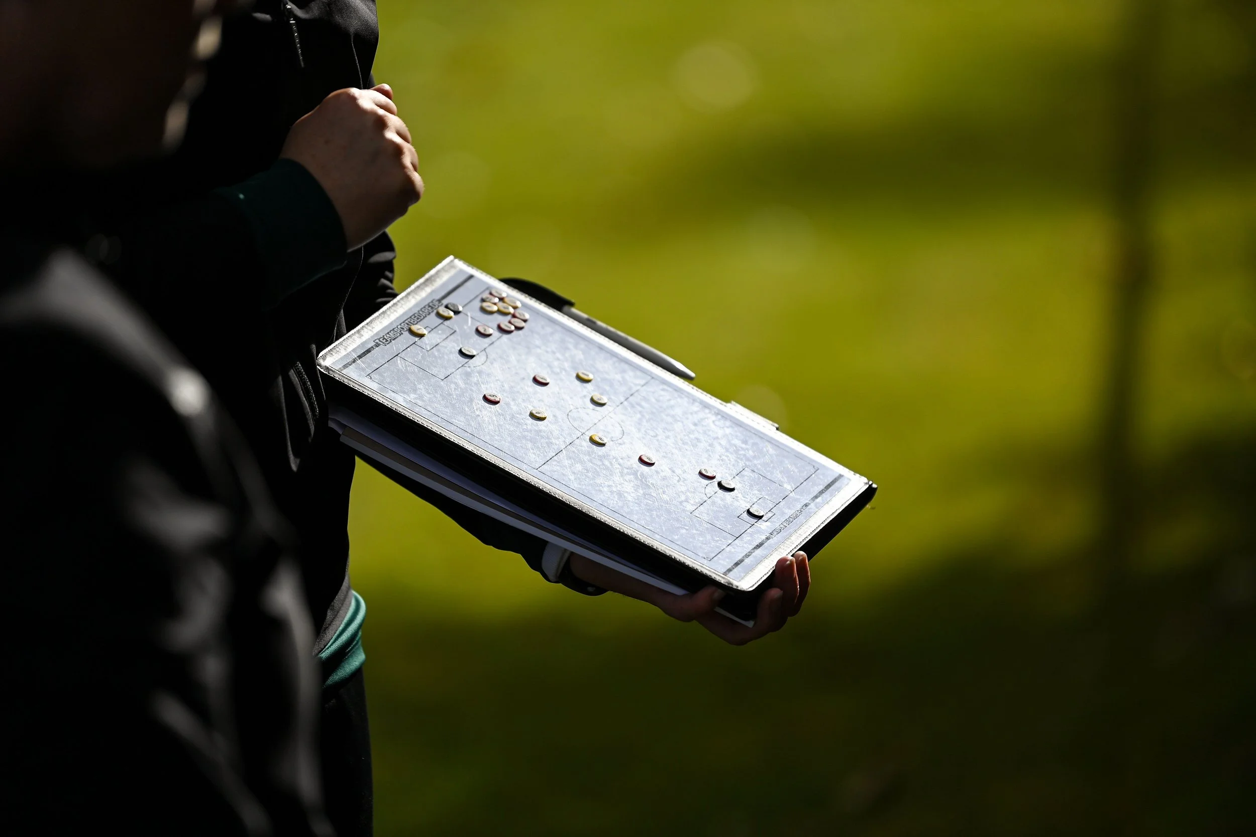 Person holding a clipboard with a soccer strategy diagram and coins, outdoors on a sunny day.