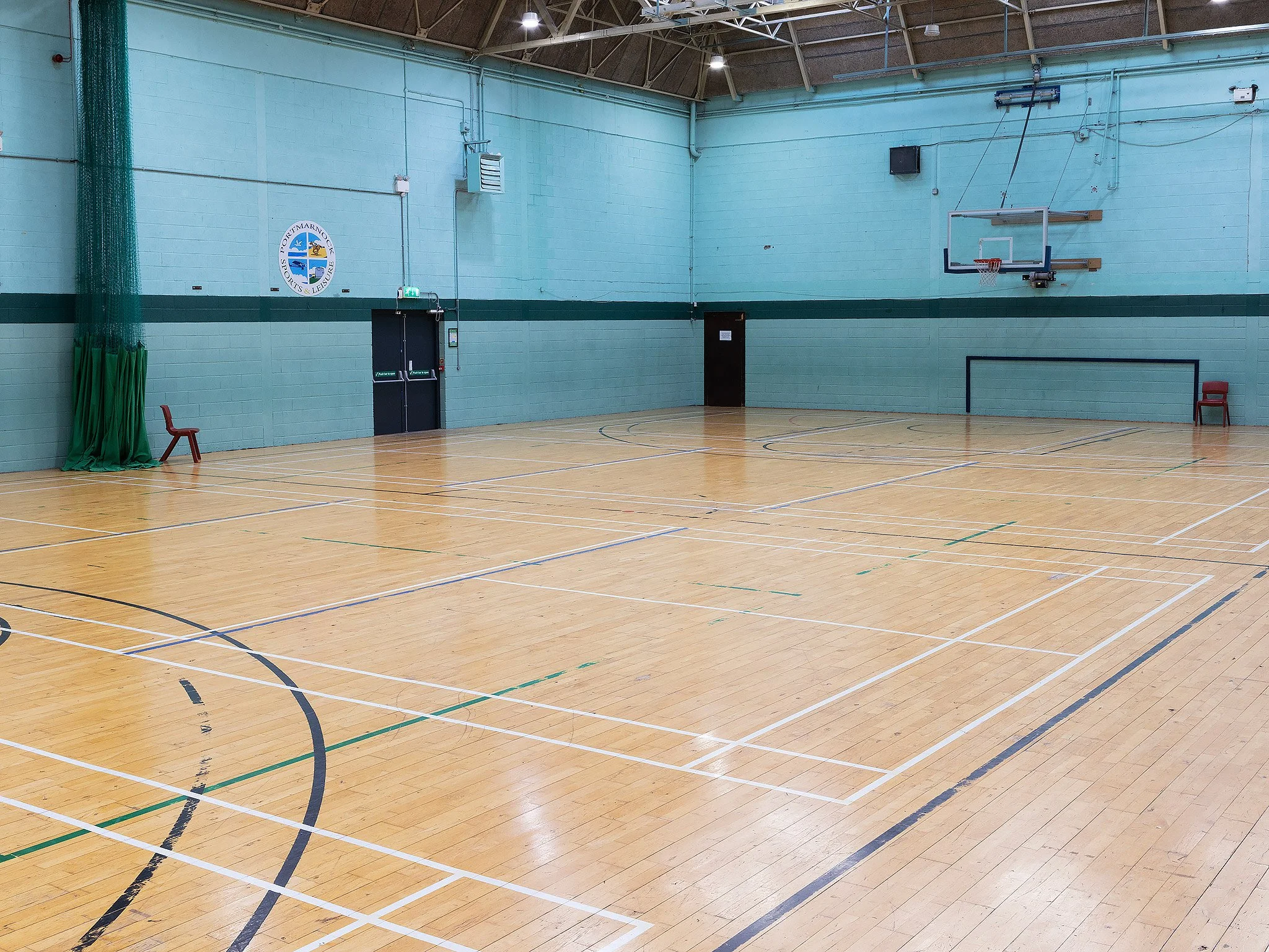 Empty indoor gymnasium with light wooden floor, teal walls, and basketball hoop on the wall.