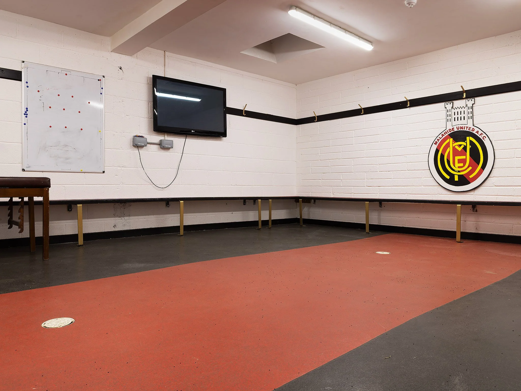 Empty locker room or team meeting area with a Malahide United AFC logo on the wall, a TV, a whiteboard, and benches.