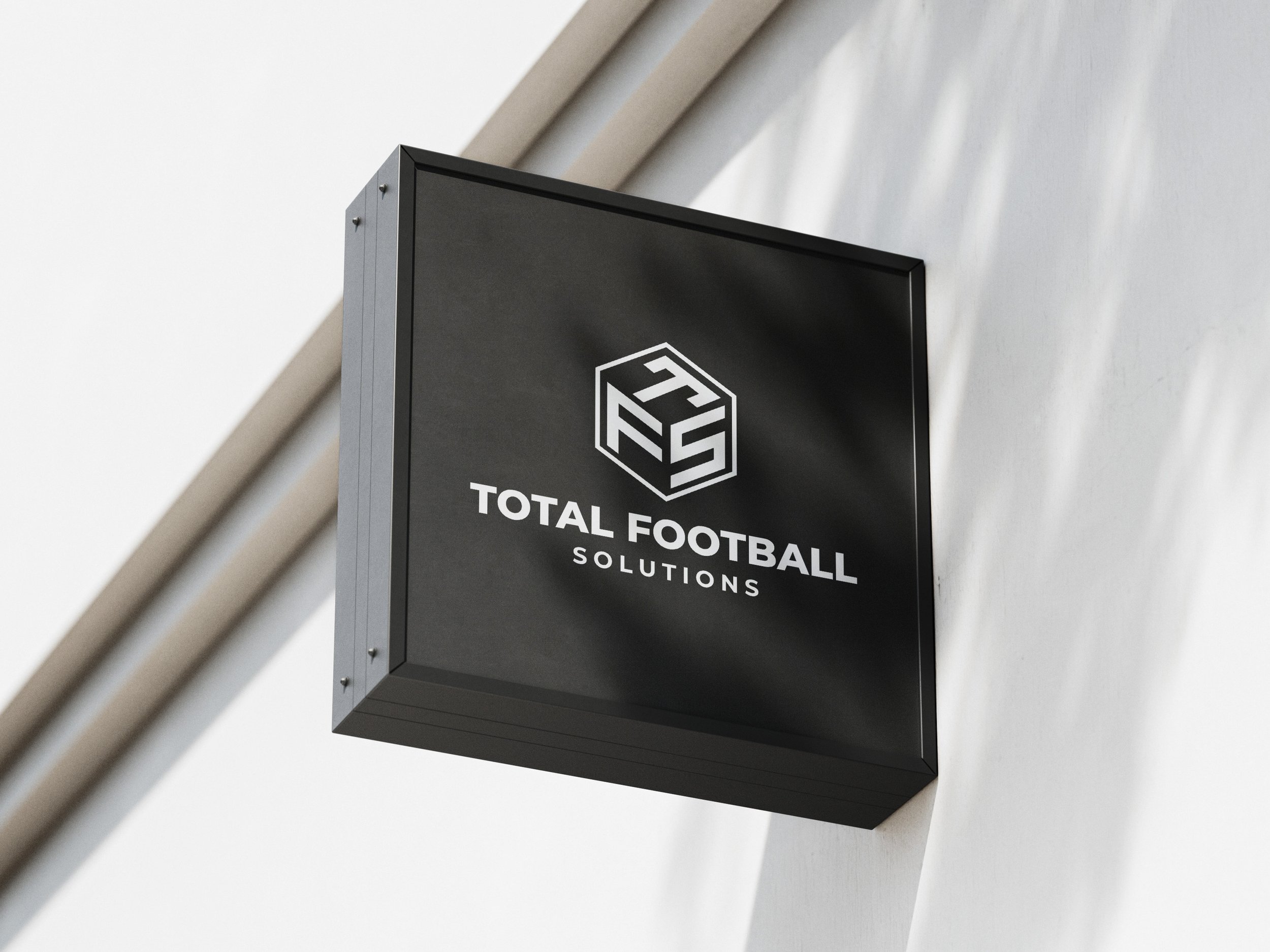 Black square sign with white logo and text reading 'Total Football Solutions', mounted on a white wall.