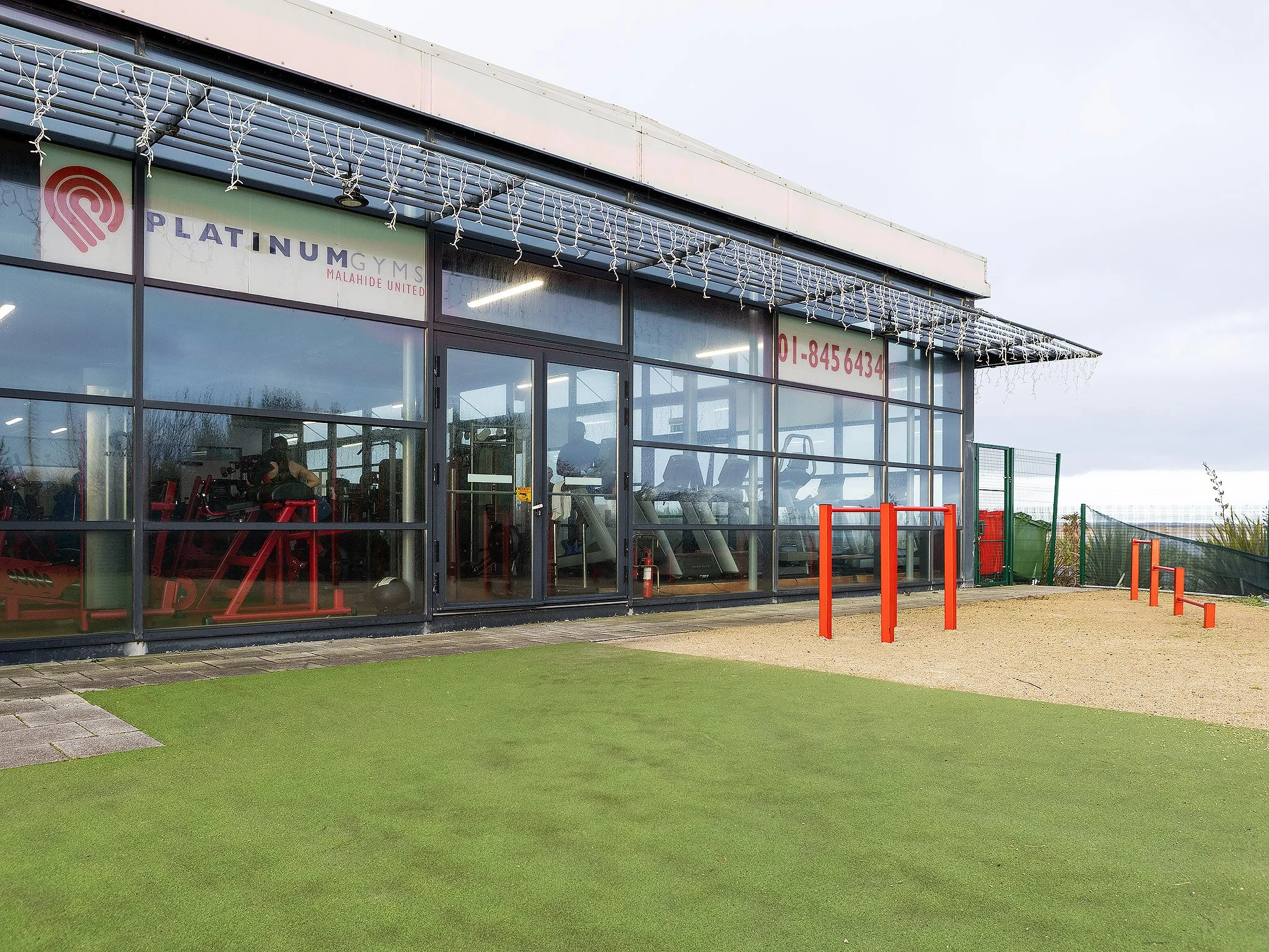 Exterior view of a glass-fronted gym called Platinum Gyms, with exercise equipment visible inside, and outdoor fitness equipment in red on a sandy and grassy area, overcast sky.