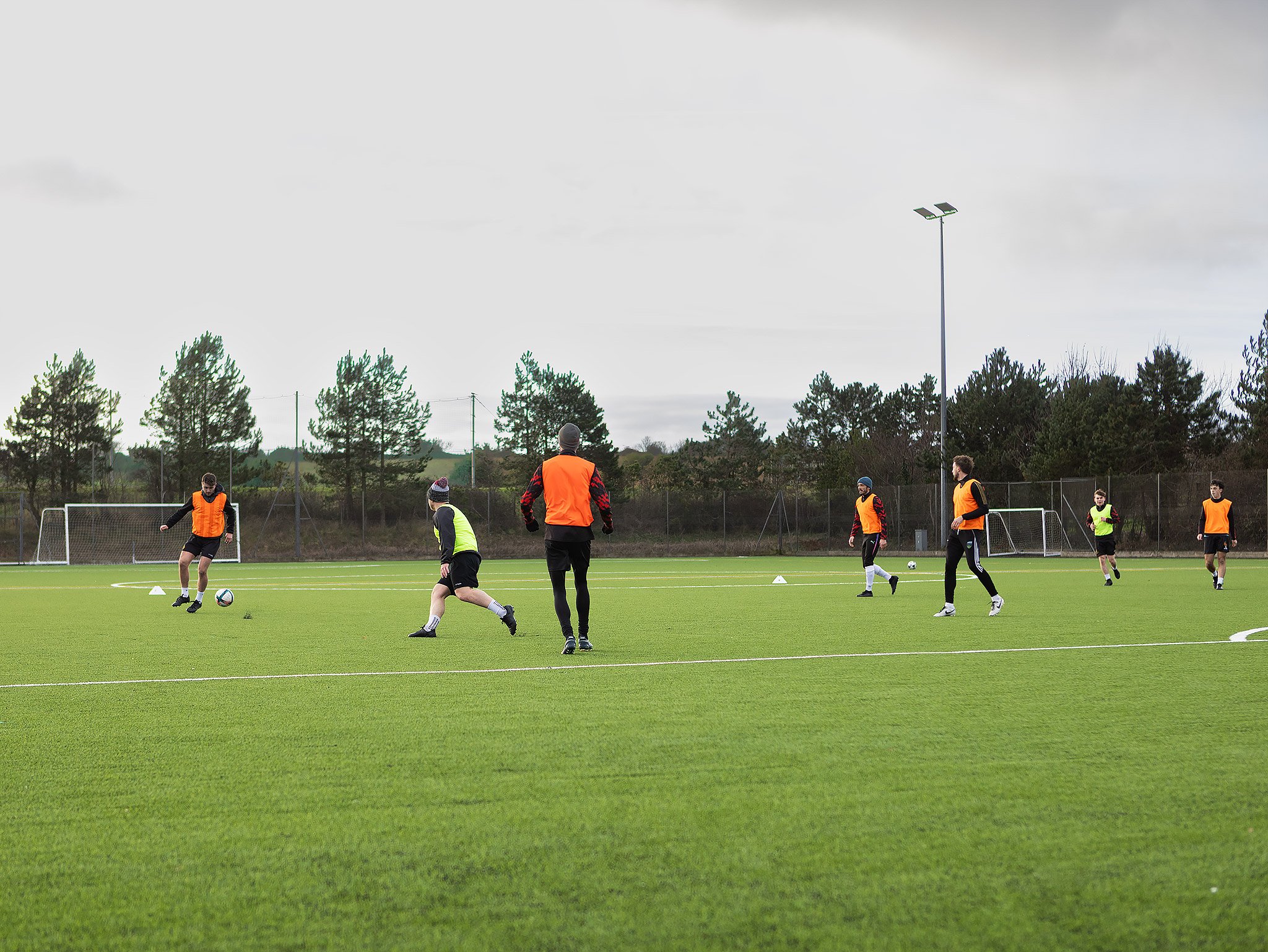 Group of young men practicing soccer on a grassy field with goalposts in the background, wearing orange and neon green training vests.