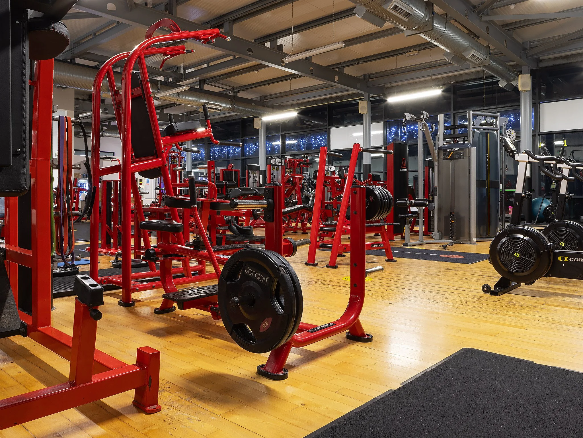 Empty gym with red workout equipment, weight machines, and cardio machines on wooden floor under industrial ceiling with ductwork.
