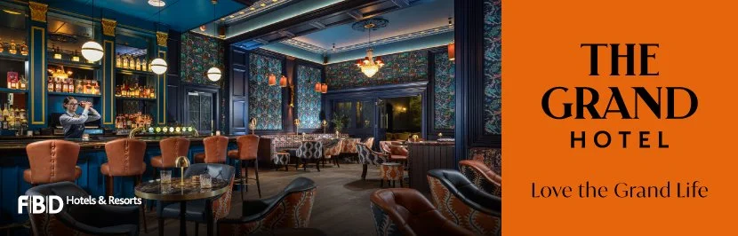 Interior of a luxurious hotel bar with blue walls, warm lighting, and orange chairs. A bartender is preparing drinks, and there are tables with elegant decor. The right side features an orange banner with black text that says "The Grand Hotel" and "Love the Grand Life."