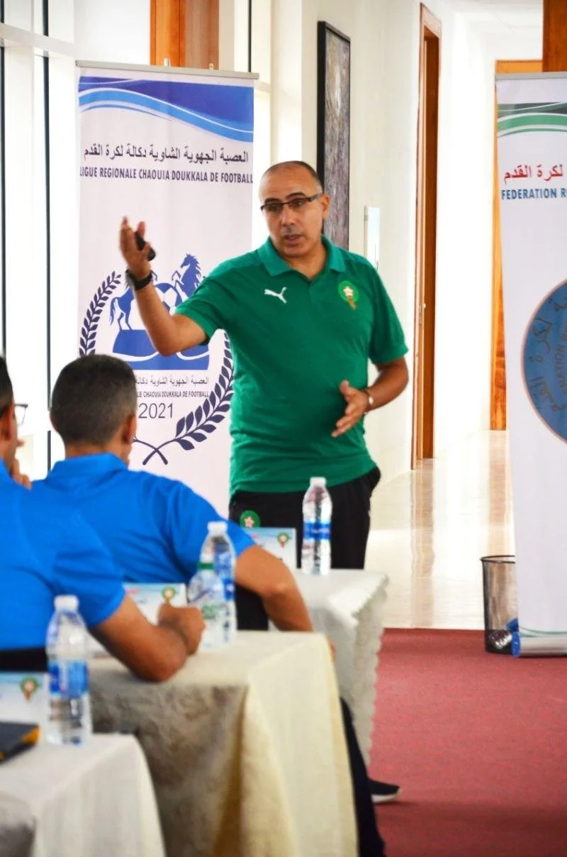 A man giving a presentation to a group of people seated at a conference table, with football banners in the background.