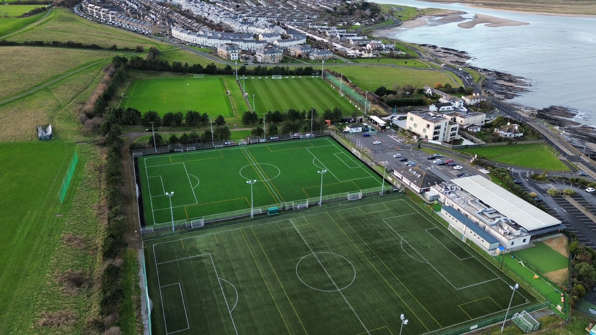 An aerial view of multiple sports fields including soccer and rugby pitches, with parking lots and residential buildings nearby, and a body of water in the background.