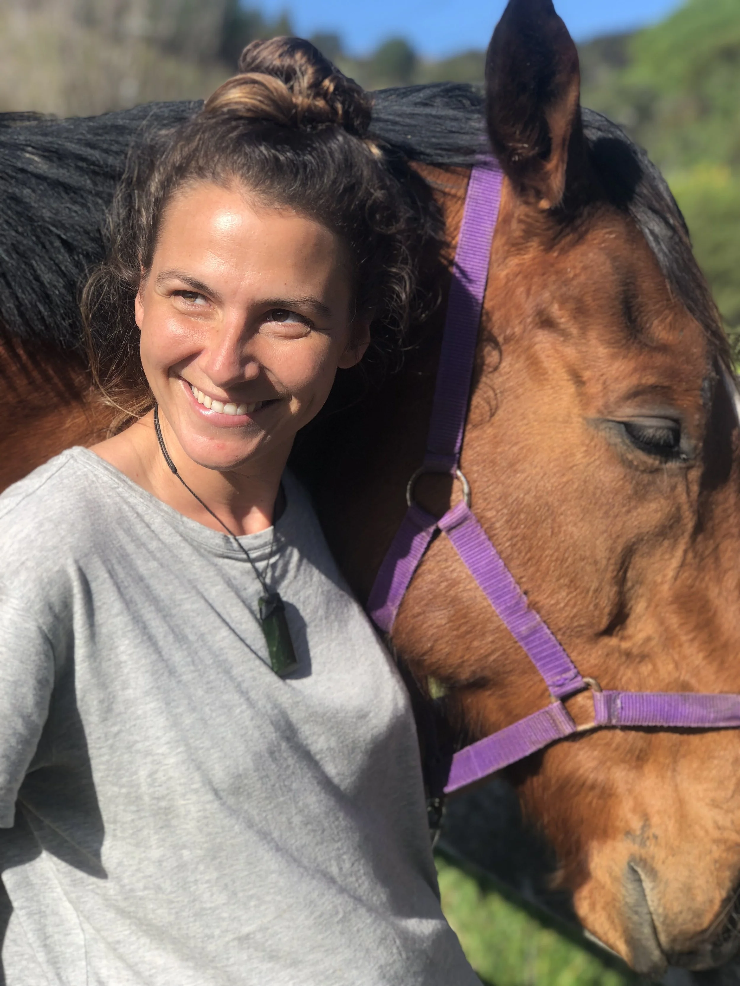 A woman smiling and hugging a brown horse with a purple halter outdoors on a sunny day. Connection course with horses north auckland