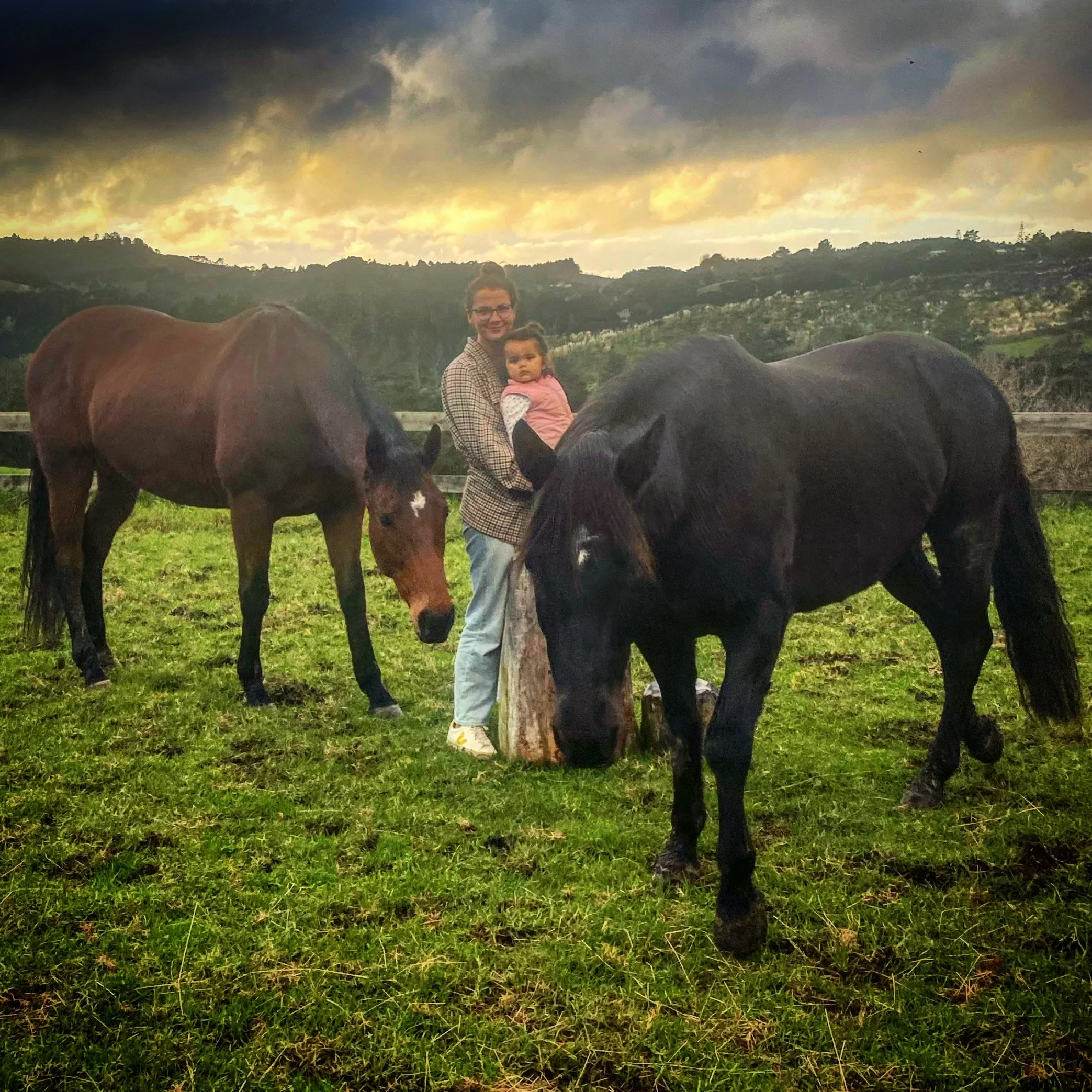 A woman holding a young girl standing between two horses on a grassy field, with a wooden fence and hills in the background during sunset. Lightness, leadership, reflection, connection, north auckland horse course for adults