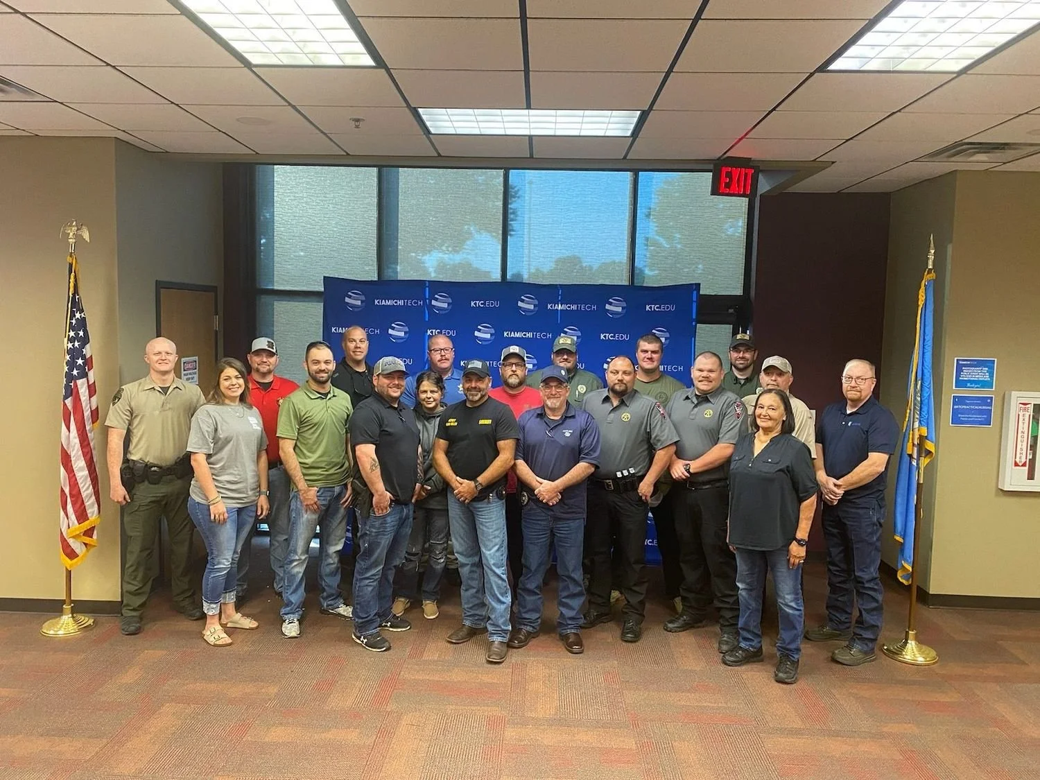 Group of people standing in front of a blue KIAMICHITECH backdrop inside a room with large windows and American and state flags on either side.