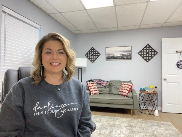 A woman with blonde hair wearing a gray sweatshirt with white text, sitting in a living room with a gray sofa, pillows, wall art, and a small table.