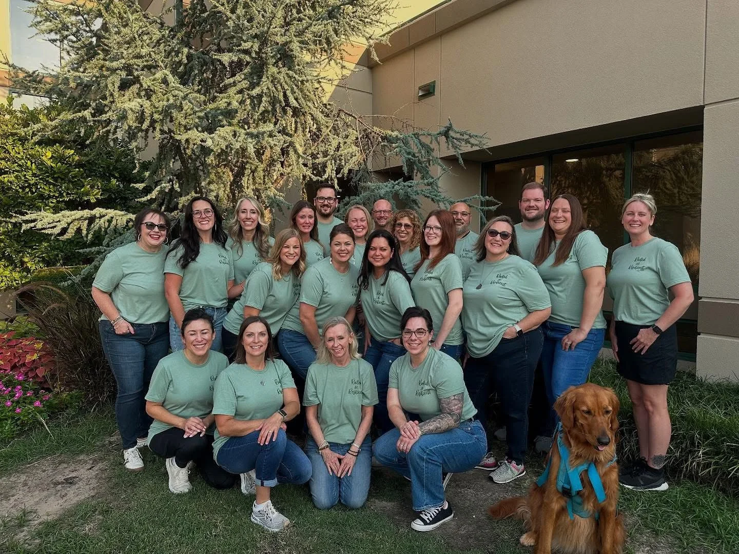 A group of 17 women and 2 men, all wearing matching light green T-shirts, pose outdoors in front of a building and trees, with a golden retriever dog sitting in front.