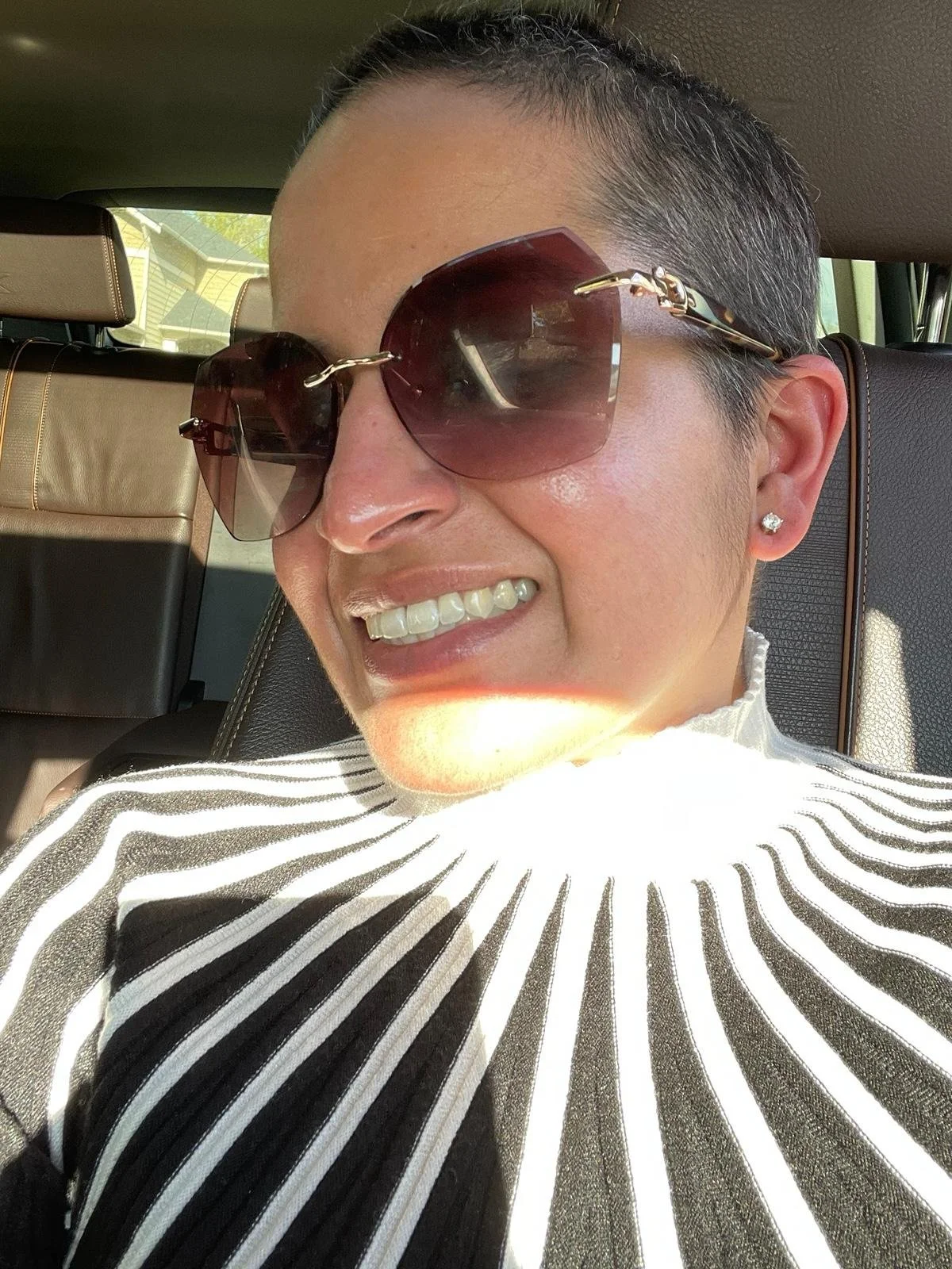 A woman wearing large sunglasses, smiling while sitting in a car with sunlight illuminating her face.