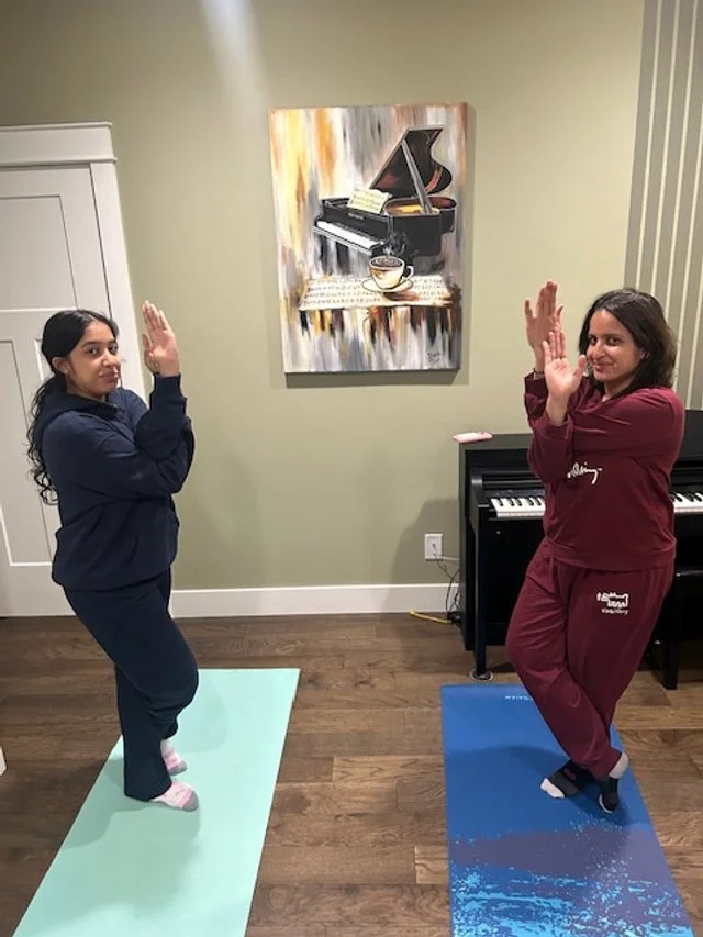 Two women practicing yoga Eagle pose (Vrikshasana) in a home sanctuary featuring a large-format piano canvas art piece, professional yoga mats, and a grand piano, representing WhiteFlutter's integration of inner and outer landscape design.