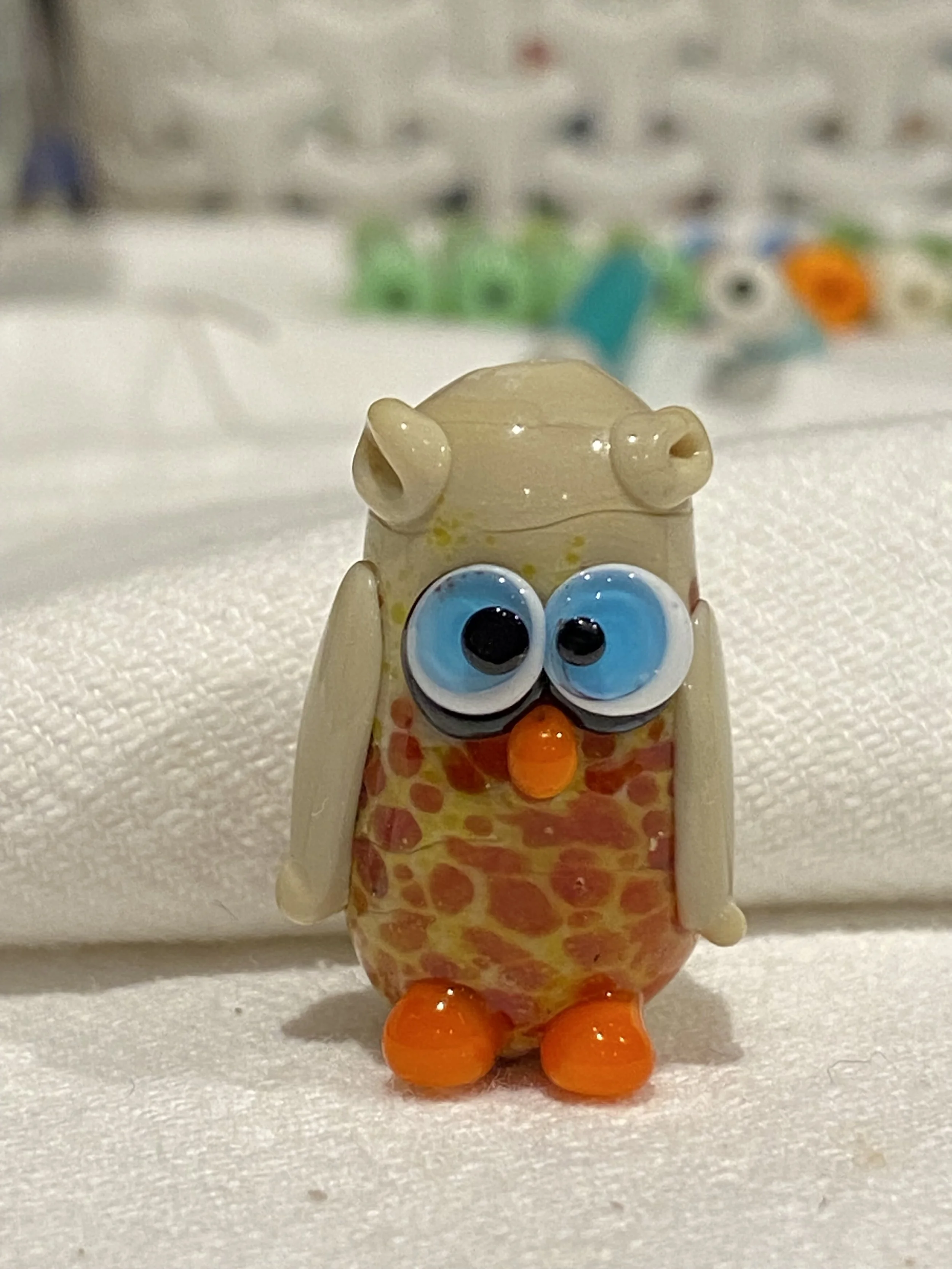 A small glass figurine of a cartoonish owl with big blue eyes, orange beak, orange feet, and a beige head and body with a giraffe-like pattern. The background shows other glass figurines in a display case.