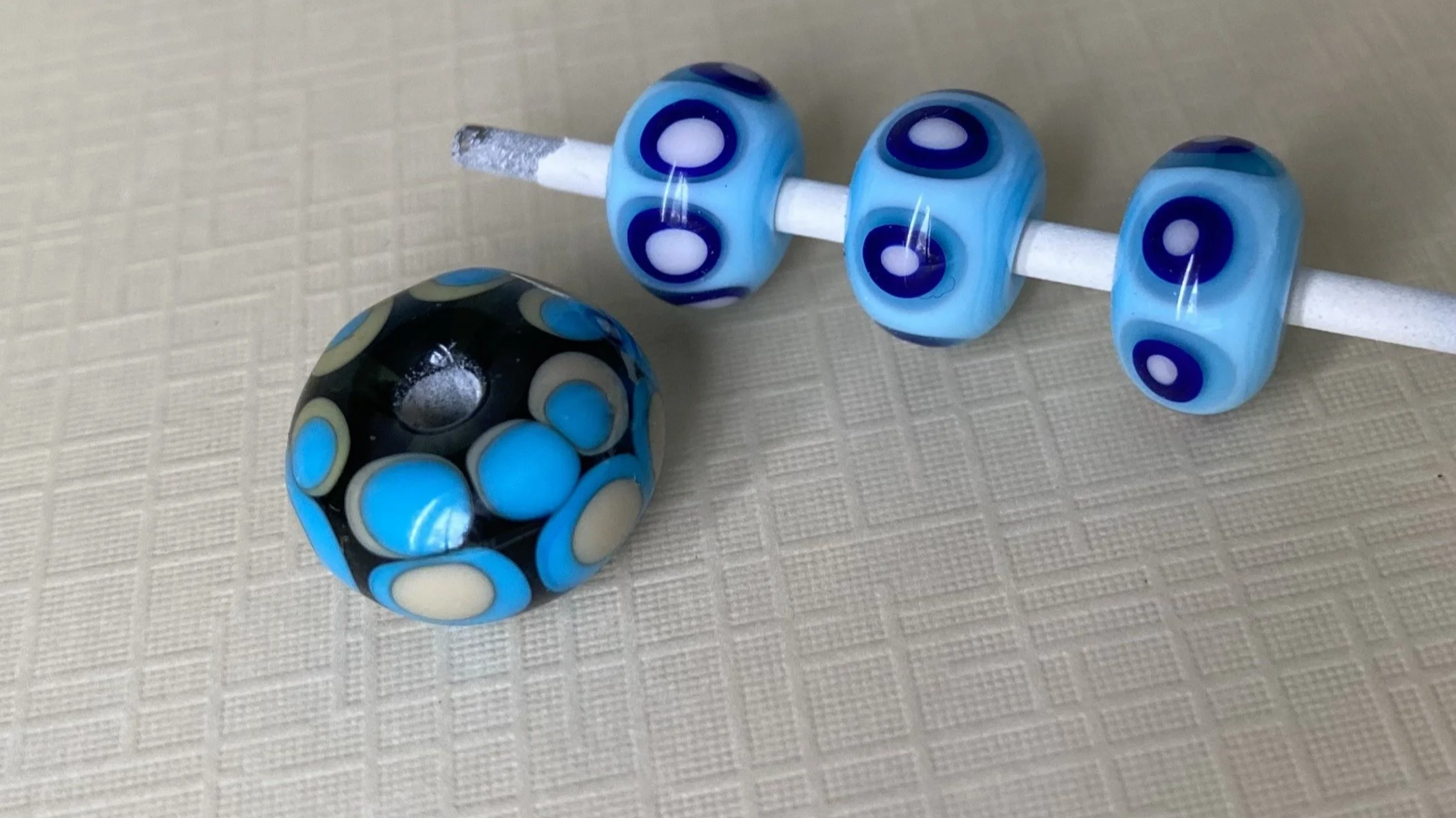 Close-up of a decorative beadwork set including a round bead with black and blue colors and a mandrel with several glass beads in blue and white patterns.