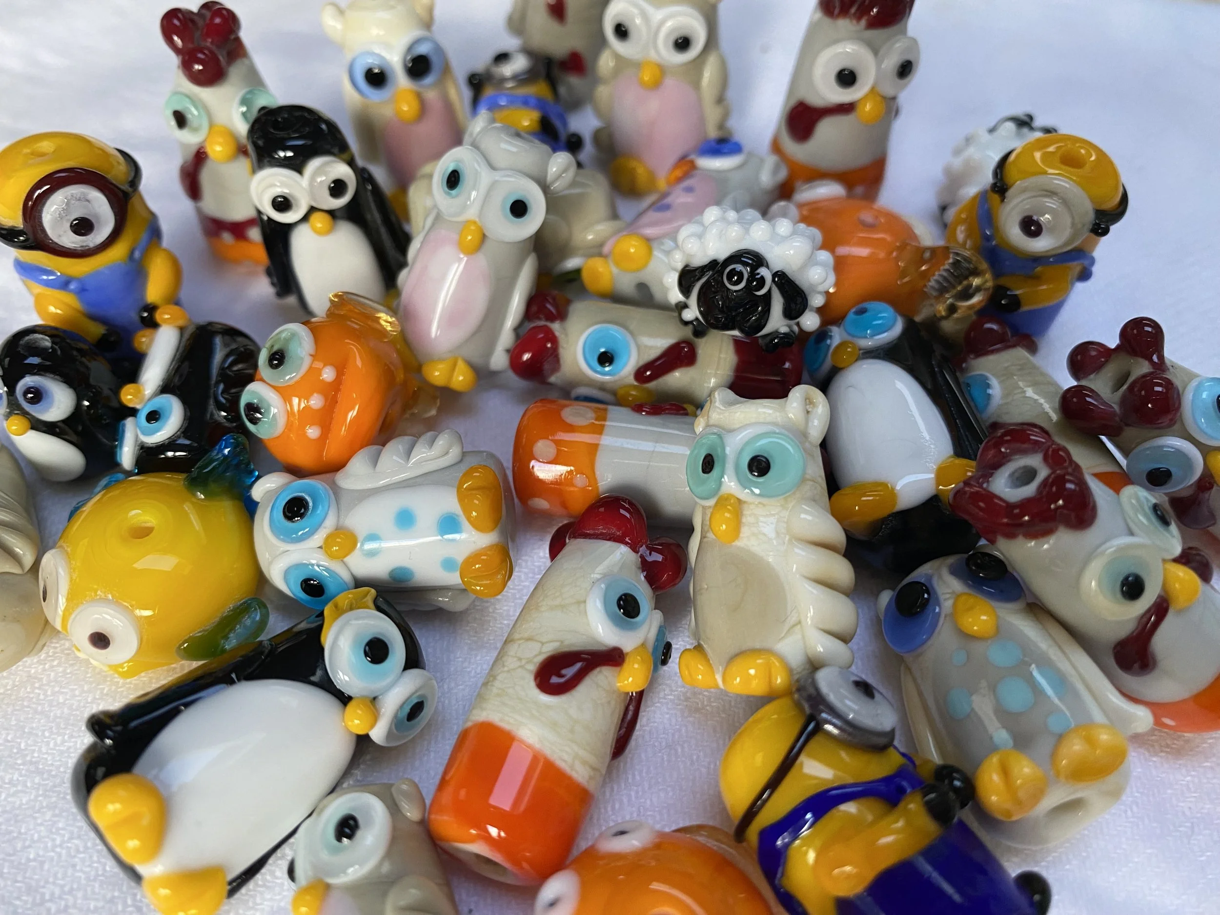 A collection of colorful glass figurines of animals, including chickens, roosters, penguins, sheep, and ducks, arranged on a white surface.