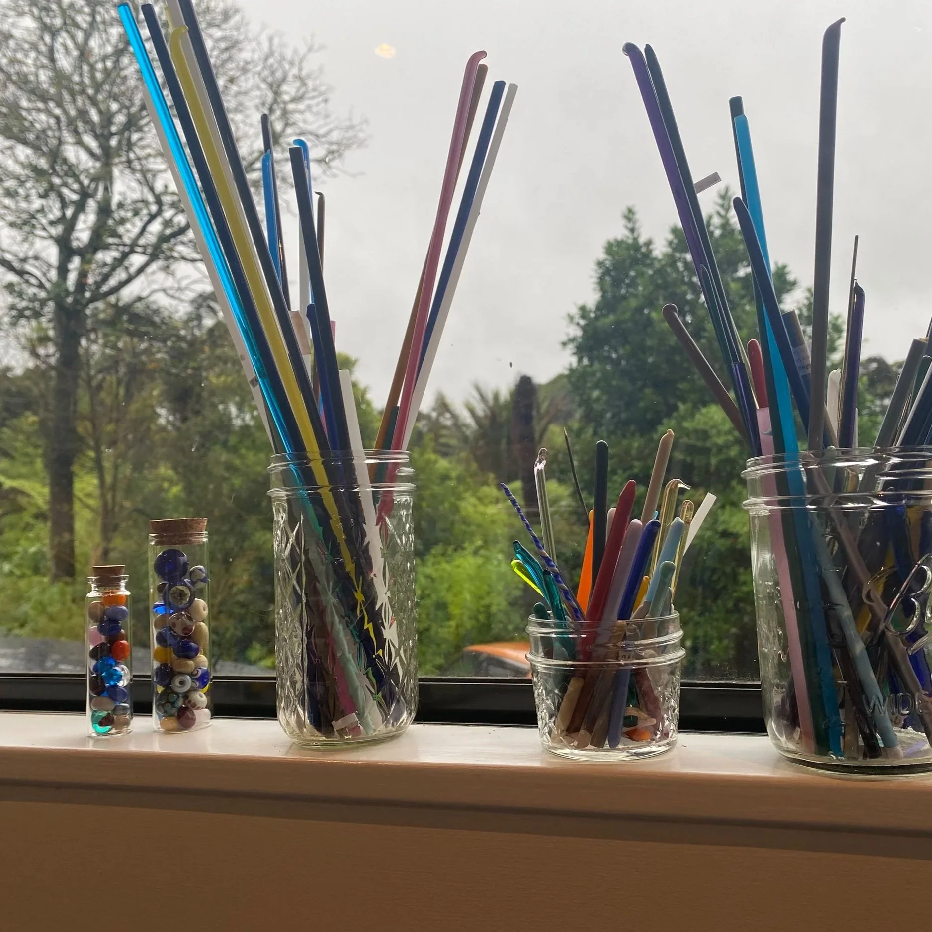Colorful glass rods in glass jars on a windowsill with trees outside