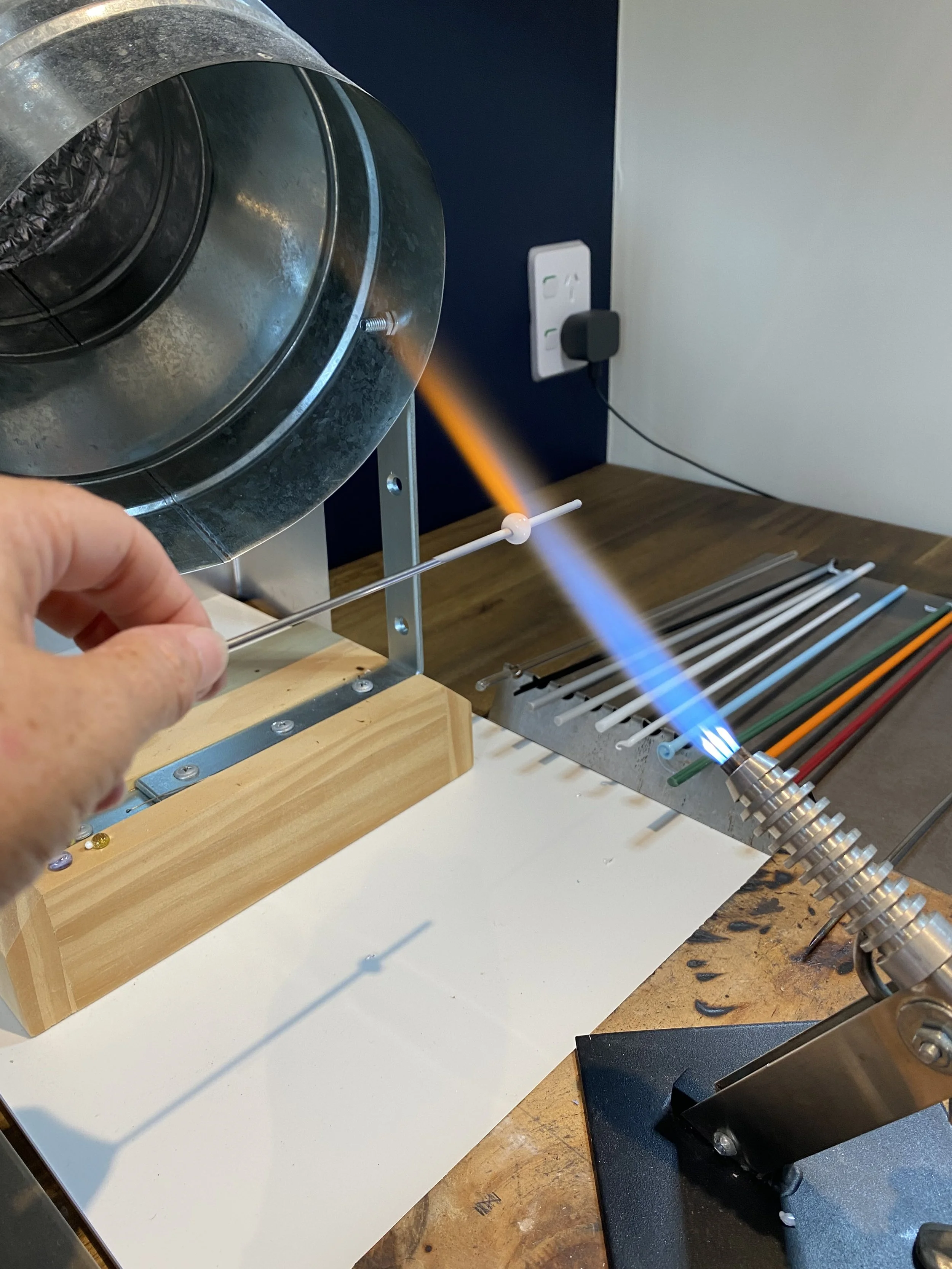A person using a blowtorch to melt glass onto a metal mandrel during lampworking process.