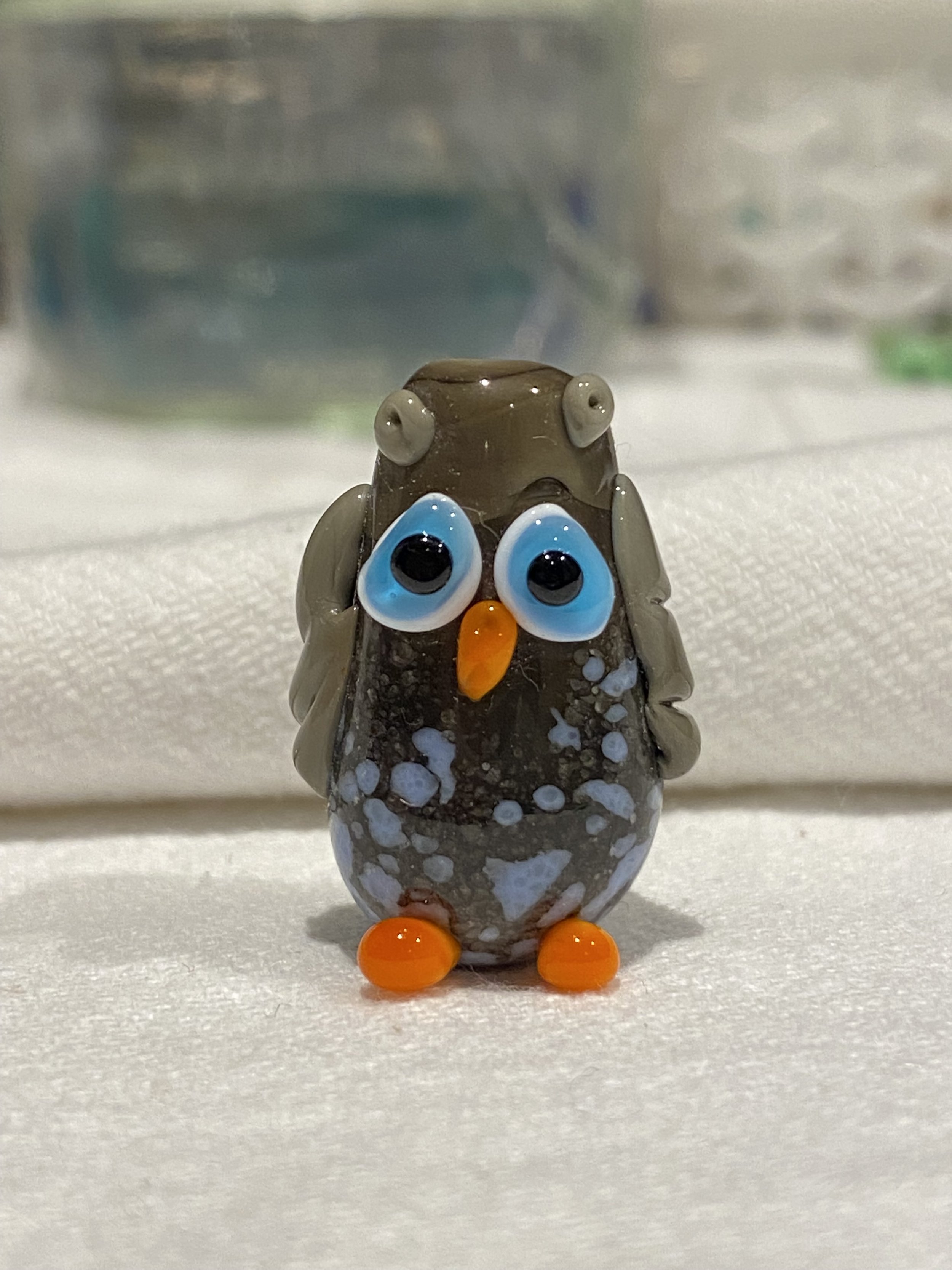 A small, glass figurine of an owl with big blue eyes, an orange beak, and orange feet, standing on a white surface.