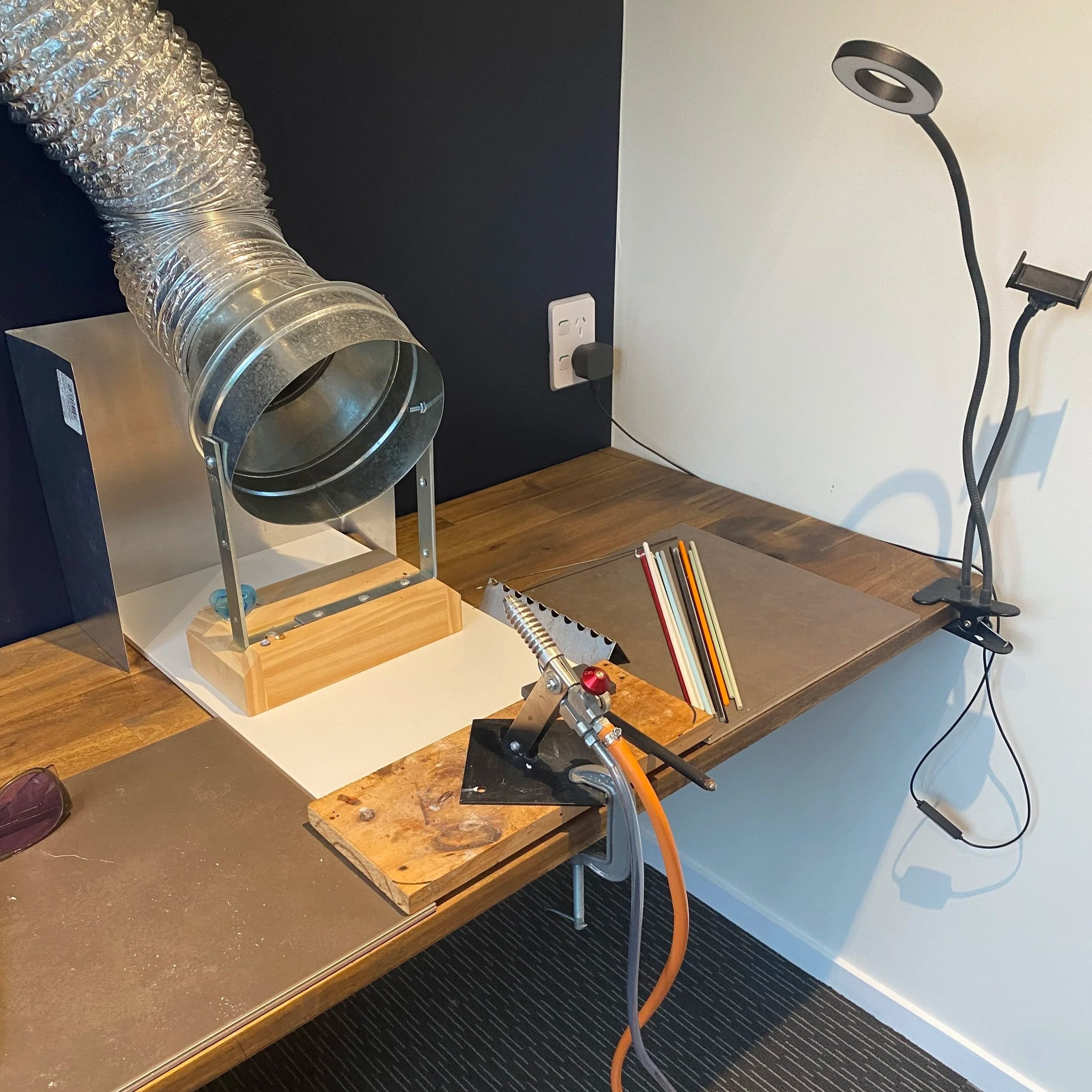 A wooden workbench with a metal hose connected to a wooden stand, a small black clamp holding an orange tube, a notebook, and three colorful sticks. A black and flexible lamp is mounted on the wall on the right side of the image, and a wall outlet with a plug is visible behind the workbench. The background features a dark and a white wall.