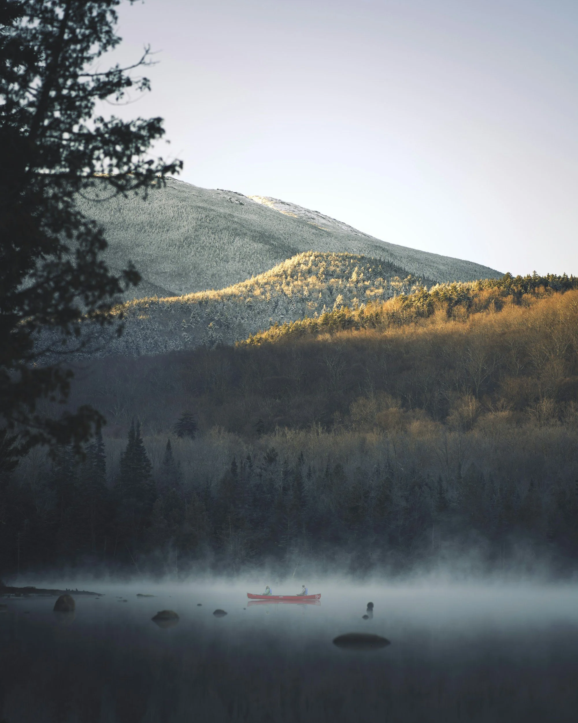 A scenic mountain landscape with a snow-capped peak, rolling forested hills, and a calm river in the foreground. There are two people in a small red boat on the river, and mist is rising from the water.