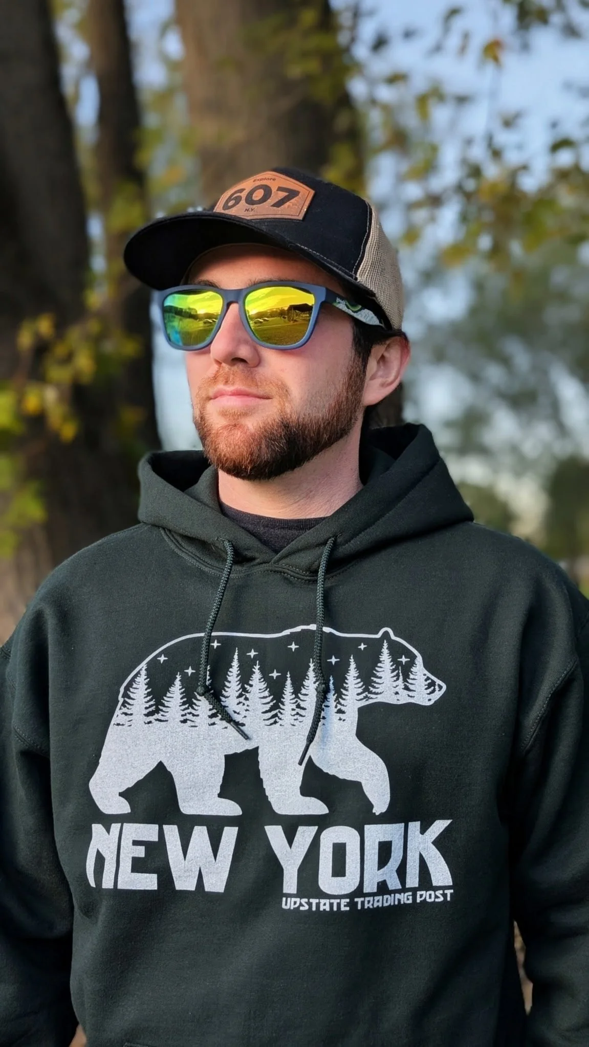 A man wearing a black hoodie with a bear and trees graphic reading "New York" and "Upstate Trading Post," reflective sunglasses, and a gray and black cap outdoors with trees and a blue sky in the background.