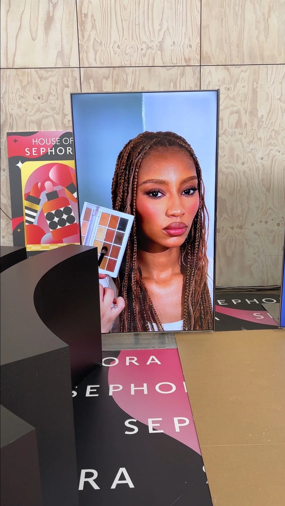 Large screen display featuring a woman with braided hair holding makeup palette, with Sephora branding and promotional posters around.