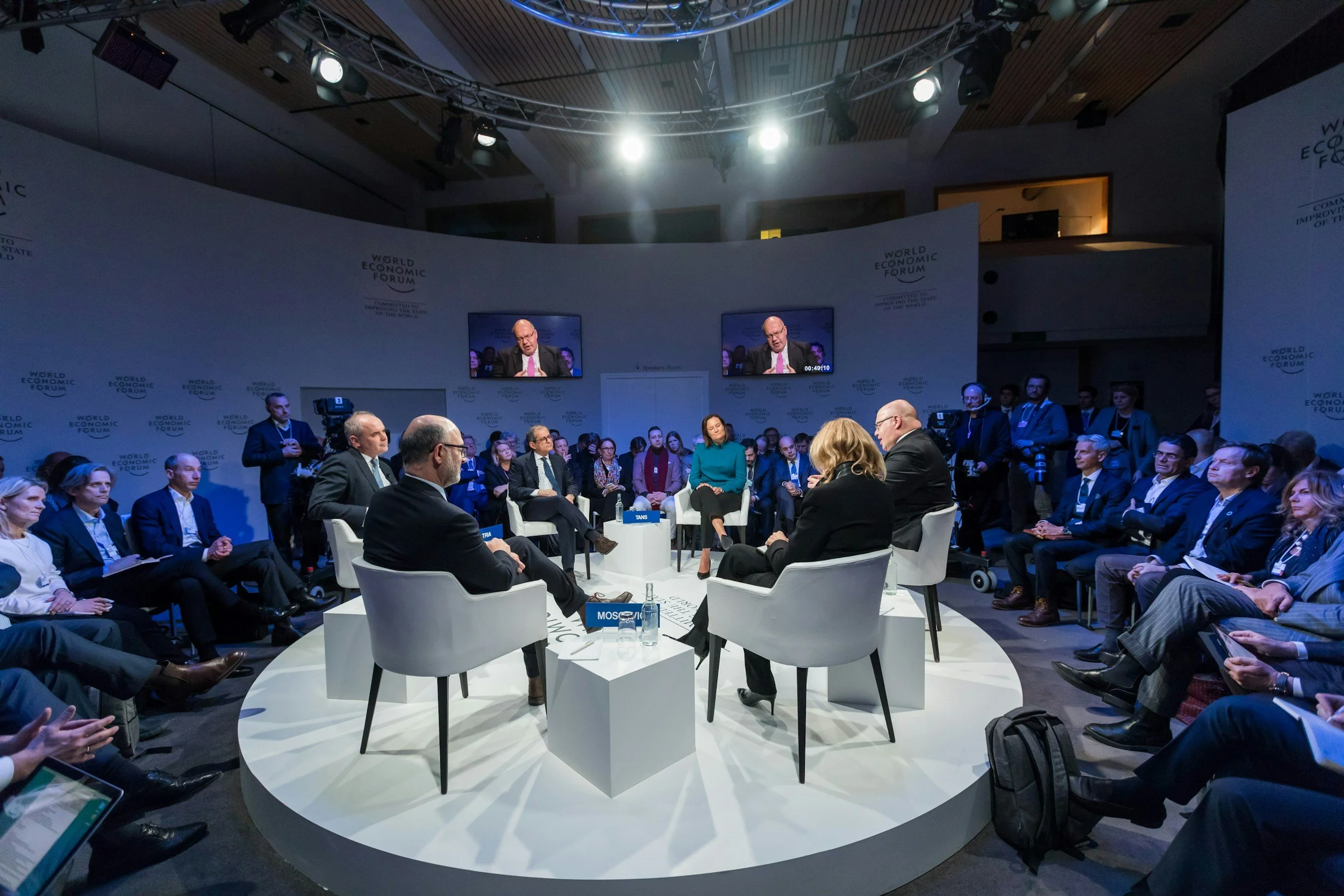 Panel discussion at the World Economic Forum with seven people seated in a circle on a stage, audience seated around, two screens displaying a speaker, and a blue logo background.