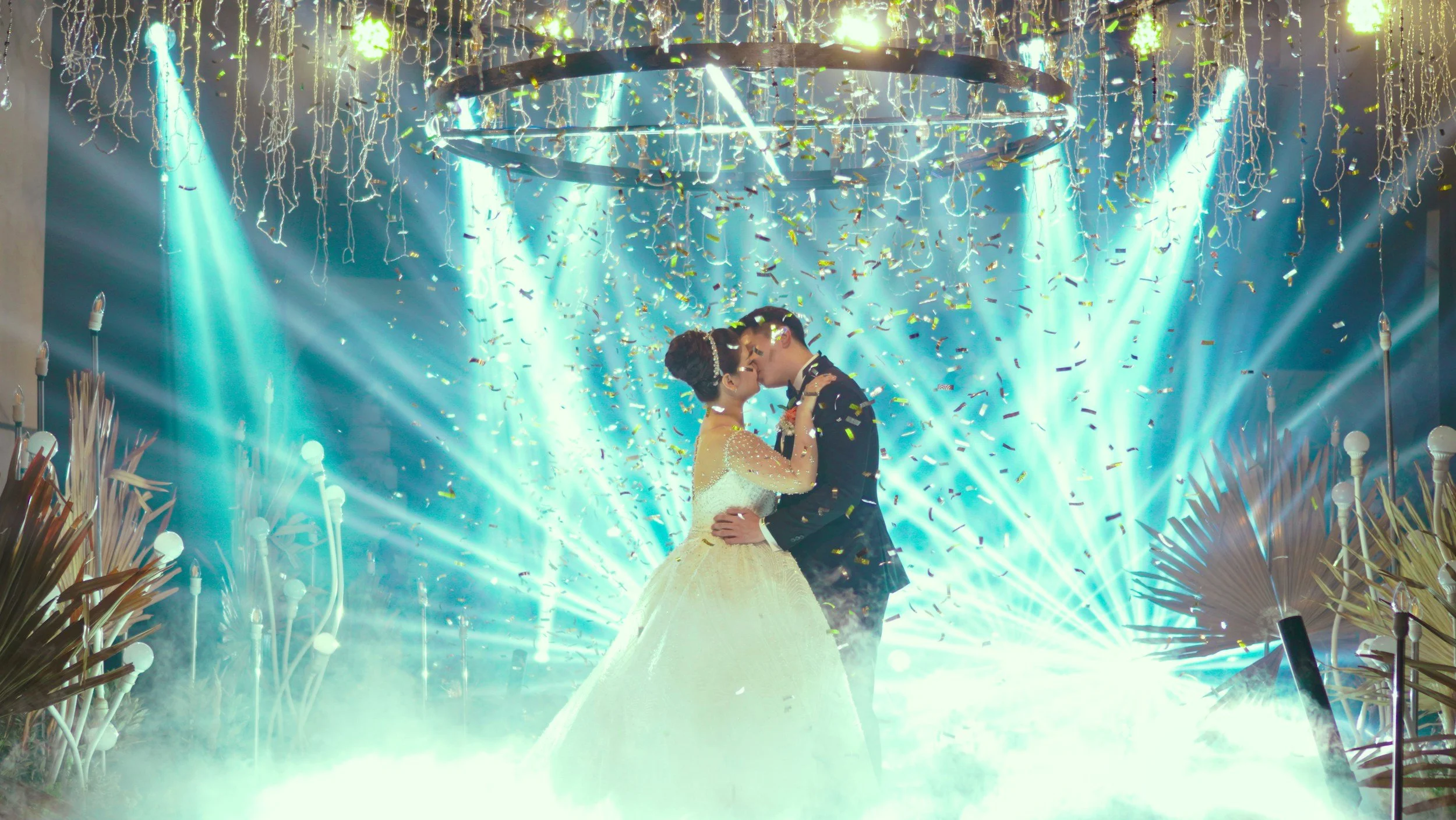 A bride and groom sharing a kiss during their wedding reception, with bright stage lights, hanging decorations, and confetti falling around them.