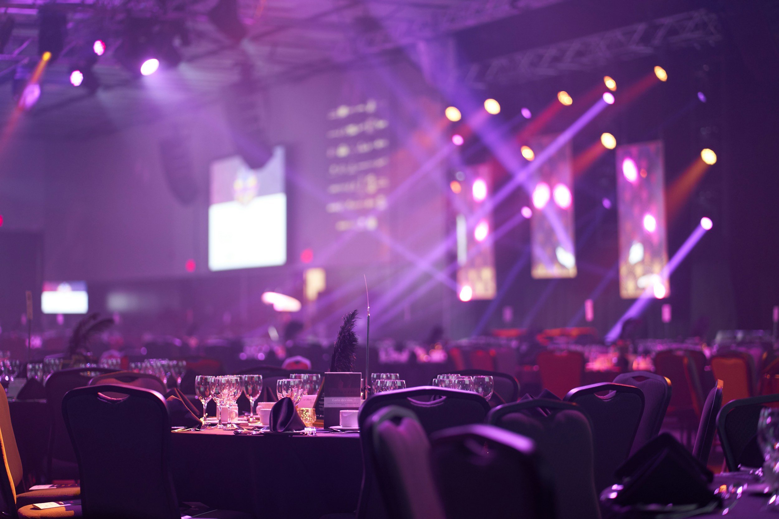 An empty event hall with round tables set with wine glasses and napkins, illuminated by purple, pink, and orange stage lighting, with a large screen and vertical light panels in the background.