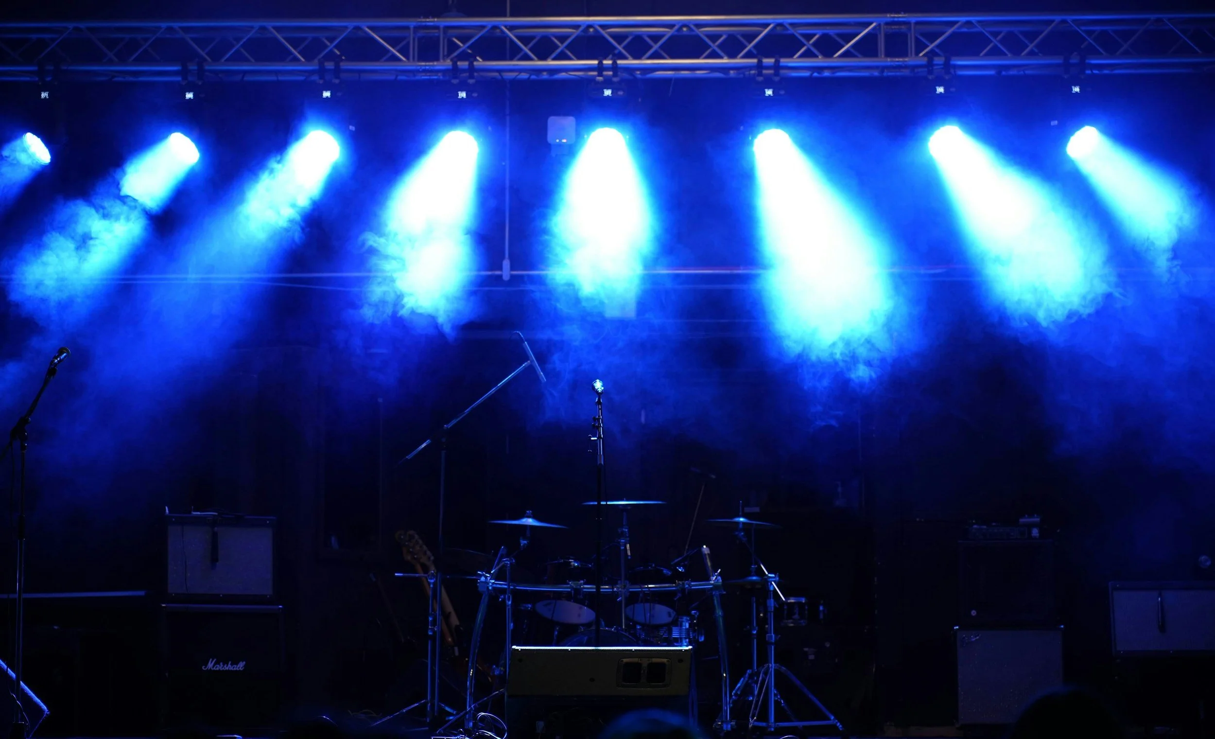 Stage with blue stage lights shining down, musical instruments including drums, guitars, and amplifiers set up, ready for a performance.