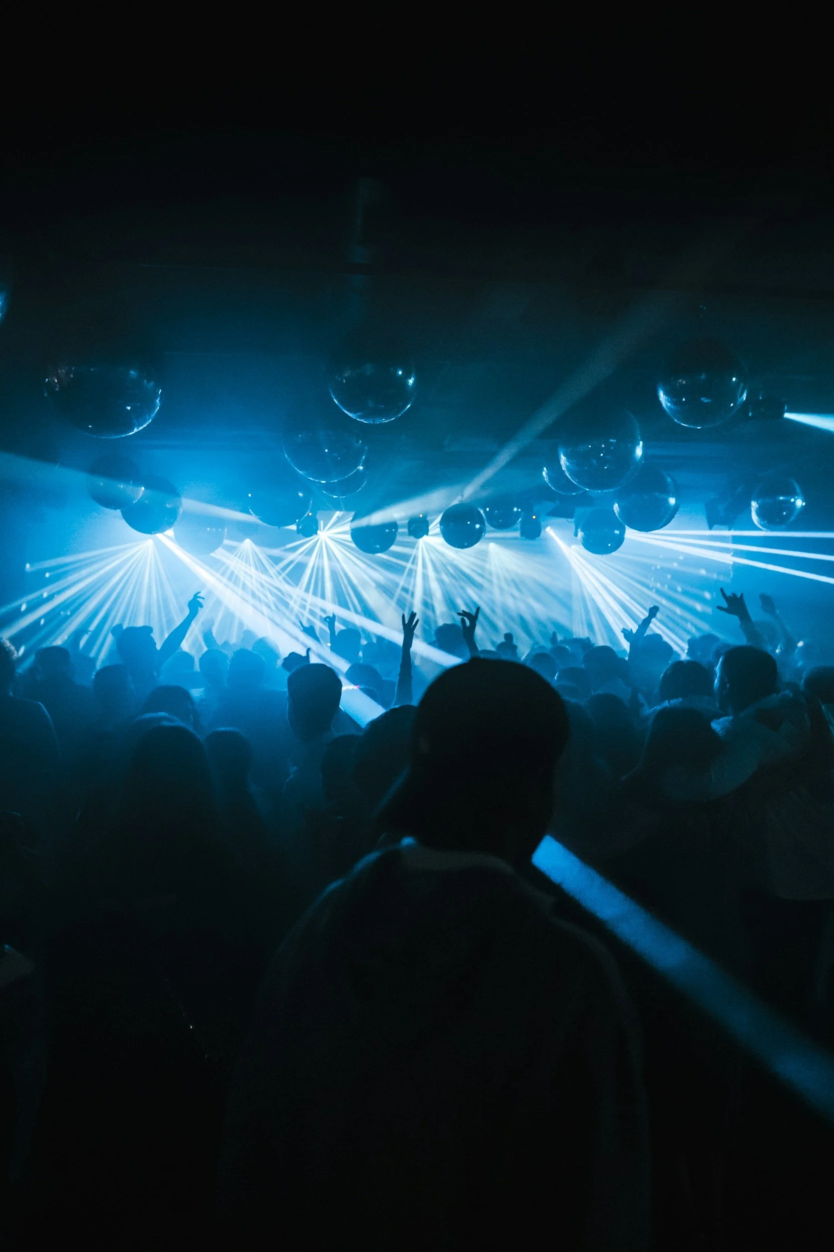 Crowd at a nightclub or concert with blue and white lights, some people raising their hands, and disco balls on the ceiling.