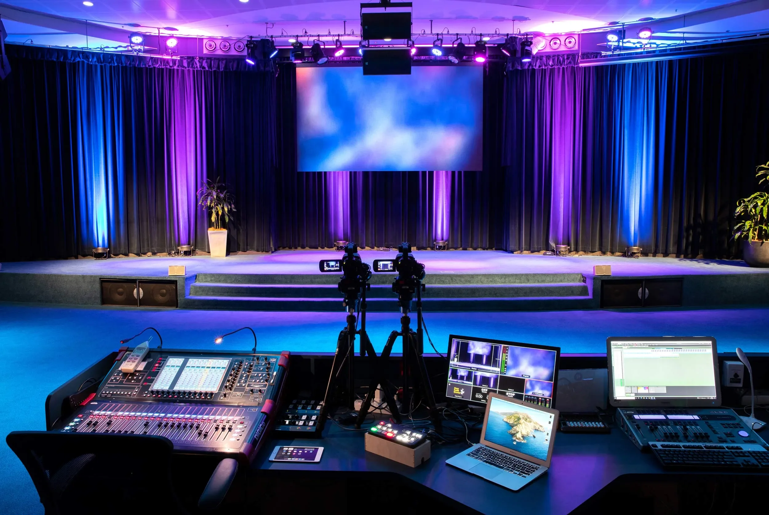 A stage set for a performance with black curtains, purple and blue lighting, large screen, potted plants, and stage lighting equipment. In the foreground, there is a control desk with sound and video equipment, two cameras on tripods, laptops, and monitors.