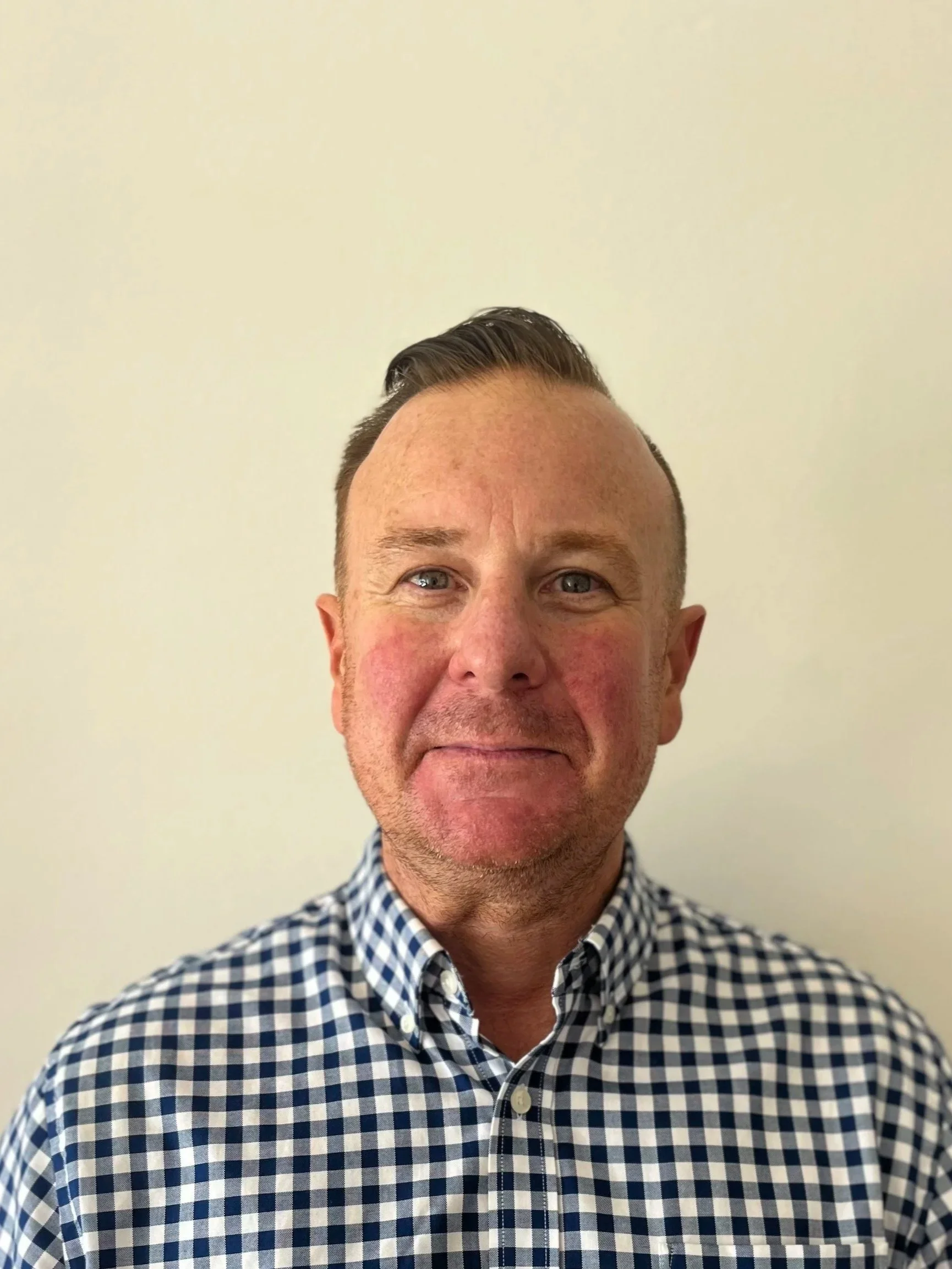 A man with short, dark hair and light skin, wearing a blue and white checkered shirt, standing in front of a plain white wall.