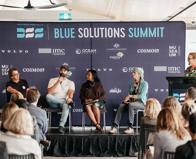 Panel discussion at the Blue Solutions Summit with four speakers on stage and an audience watching, backlit by a large banner displaying event branding and sponsor logos.