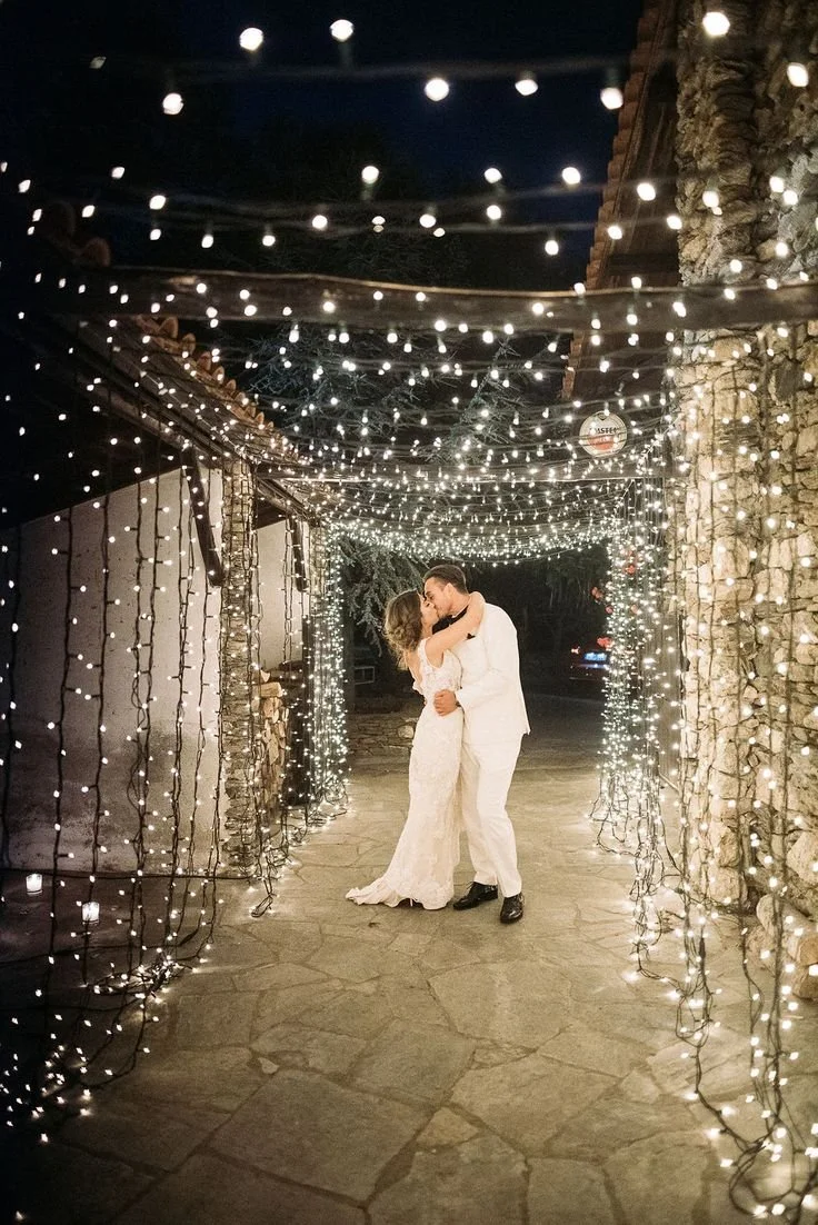 A couple in wedding attire sharing a kiss under a canopy of string lights at night.